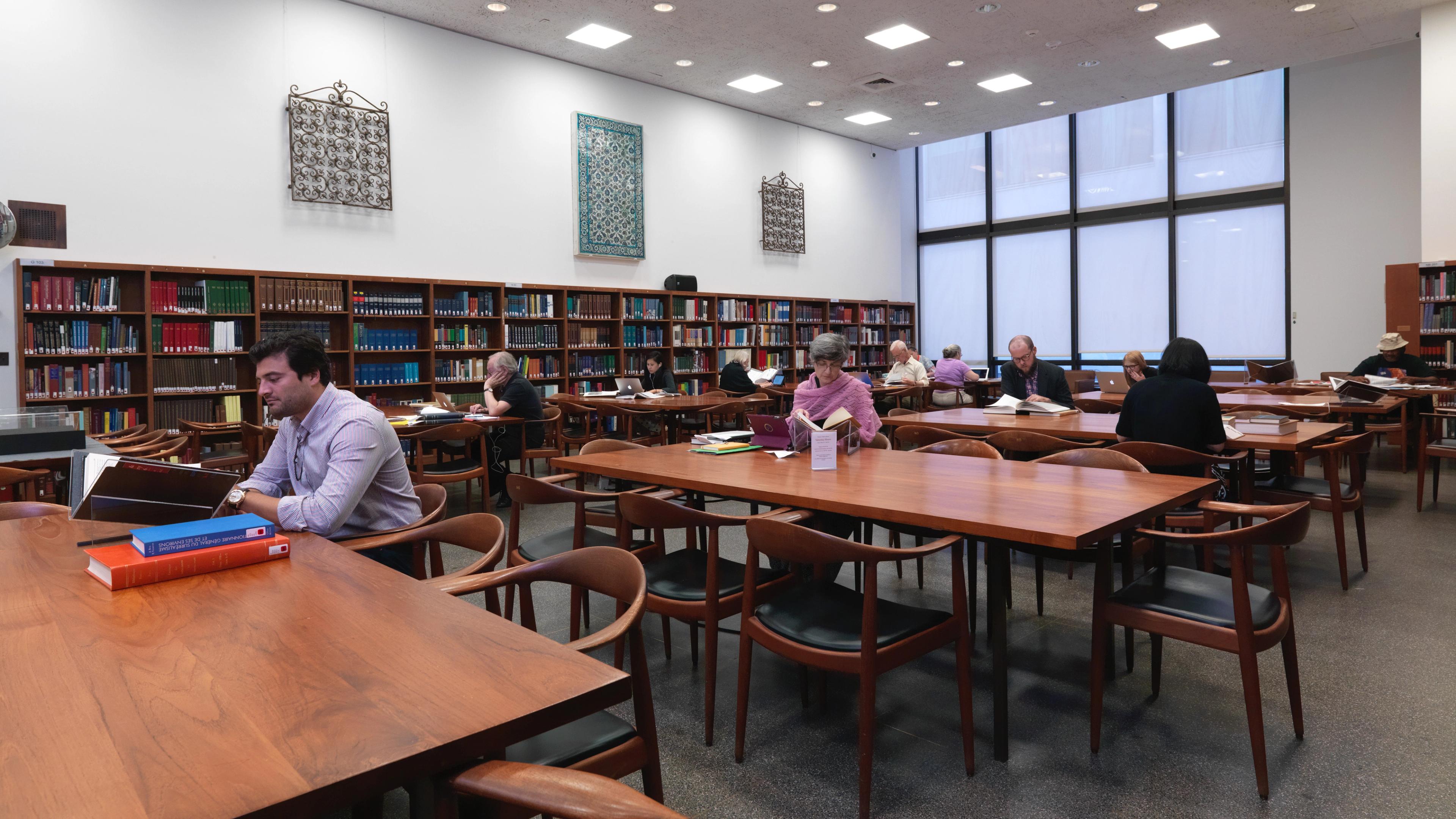A spacious library with wooden tables, shelves filled with books along the back wall, and floor-to-ceiling windows on the right wall. Several individuals are reading books at the tables.