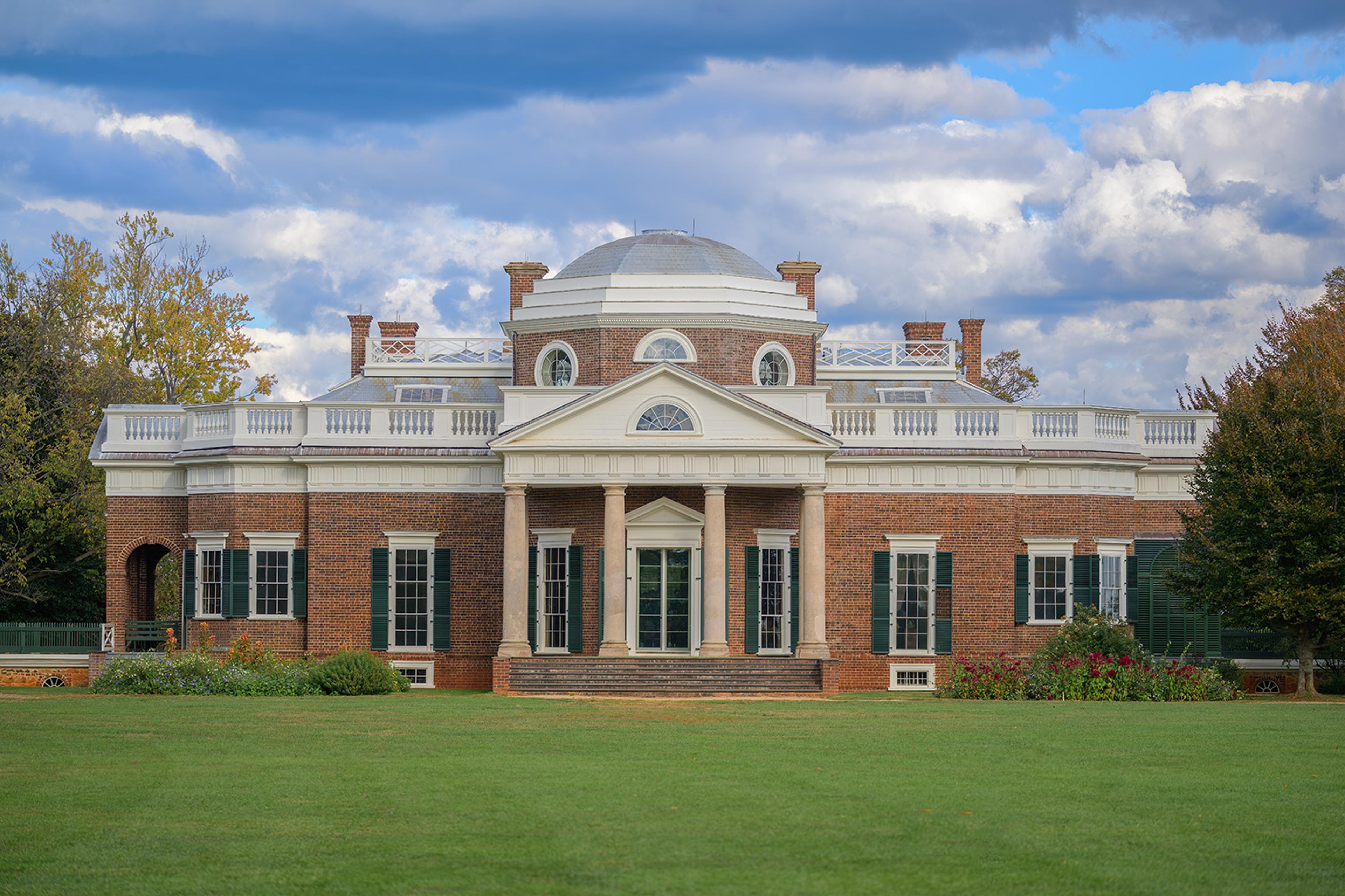 Historic red-brick neoclassical mansion with white columns, balustrade, and domed roof, fronted by a green lawn beneath dramatic clouds.