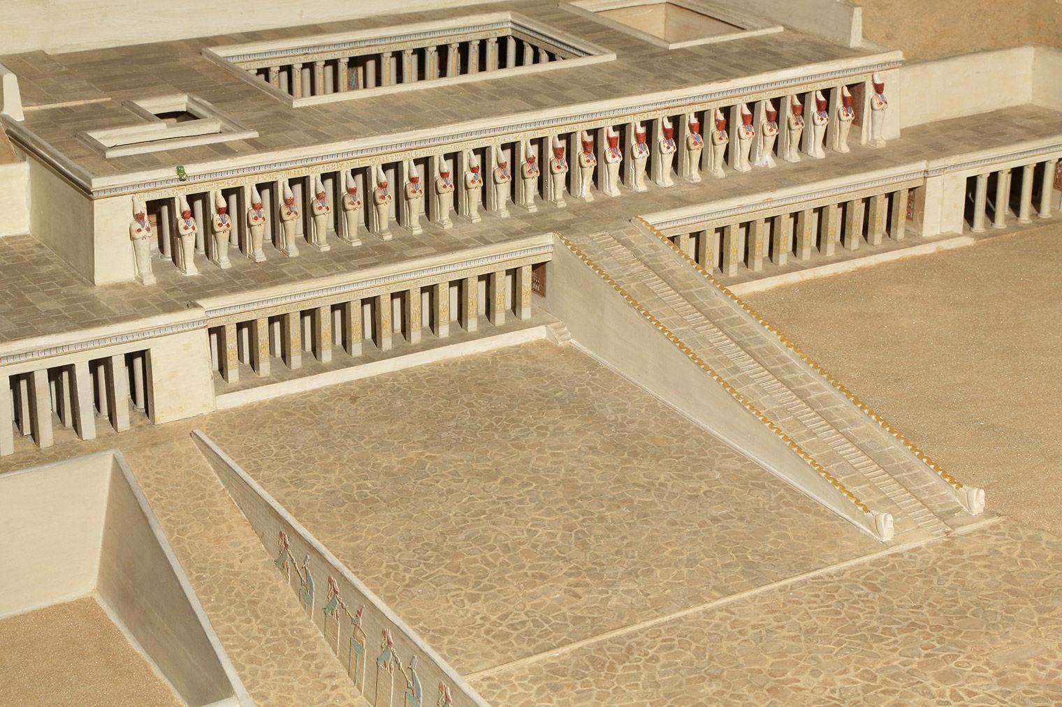 Closeup of top part of terraced temple, with ramp leading up to portico with mummiform statues in red and white crowns.