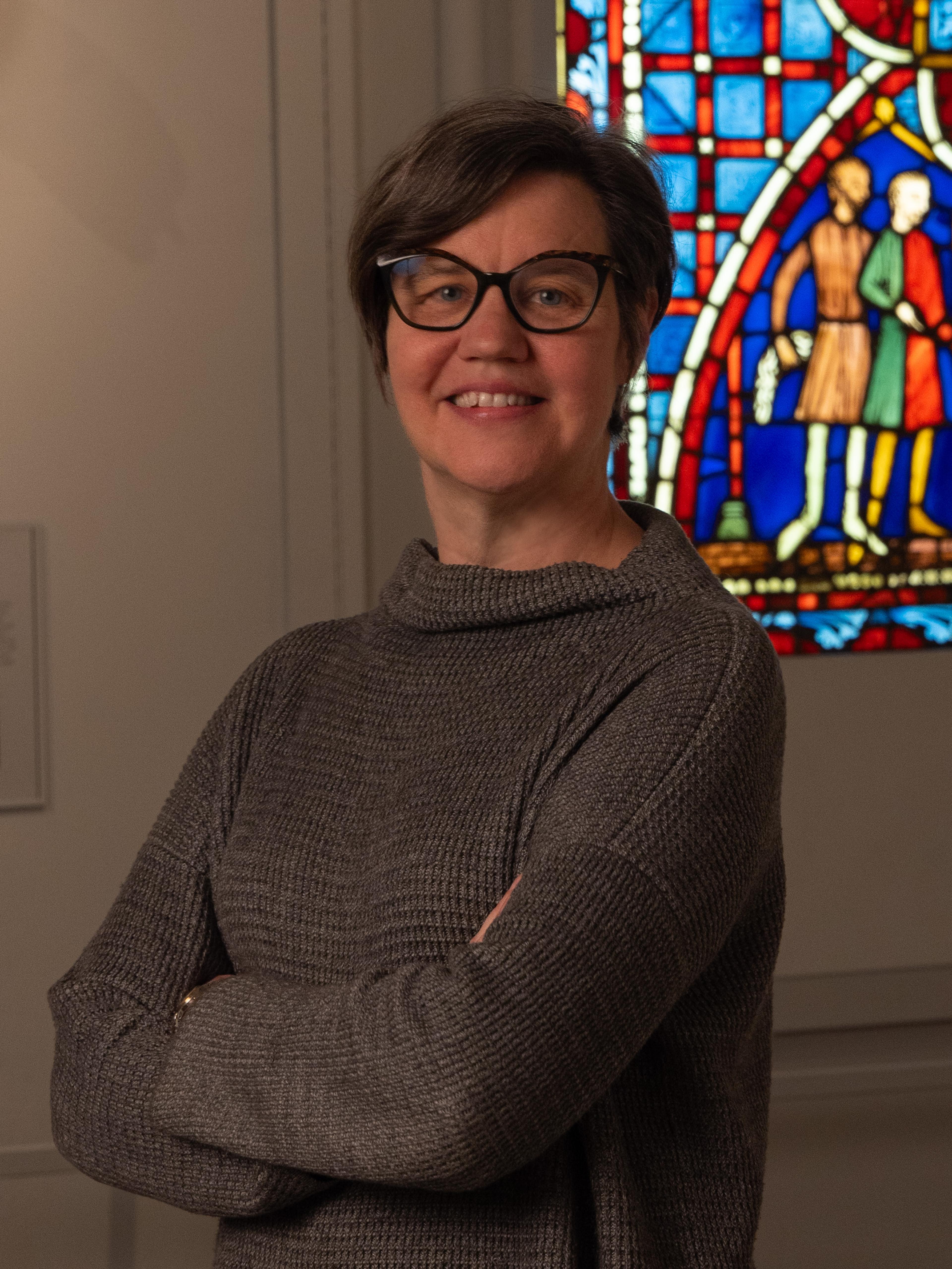 A woman with short hair crosses her arms and smiles in front of a stained glass window