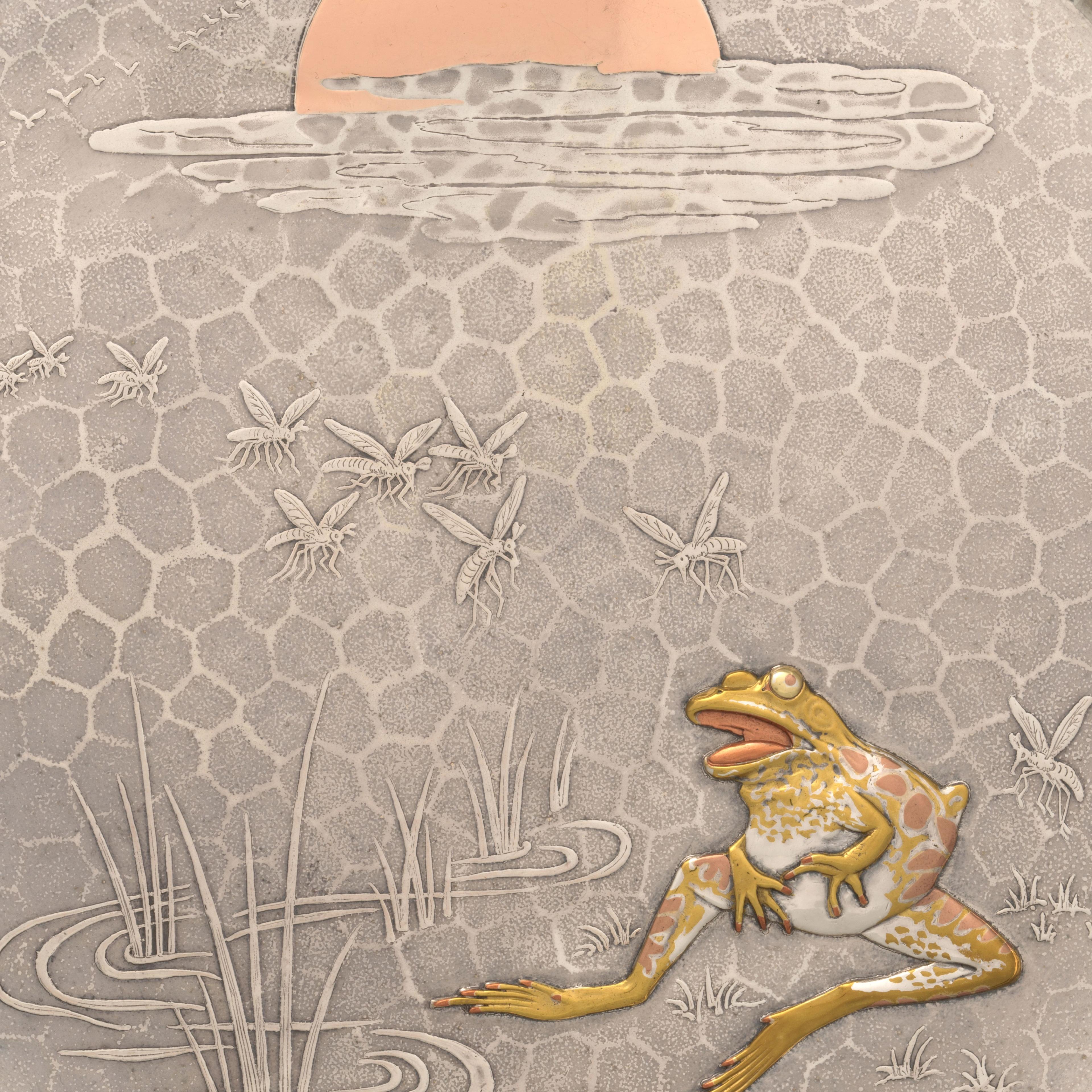 Detail of a silver tray featuring the design of a frog seated at the edge of a grassy pond with a queue of mosquitos approaching from a setting sun on the horizon. The surface has a hexagonal-shaped texture. The grass and mosquitoes protrude in a low relief on the tray surface. The front is more heavily sculpted and plated with mixed metals that are silver, gold, and copper in tone.