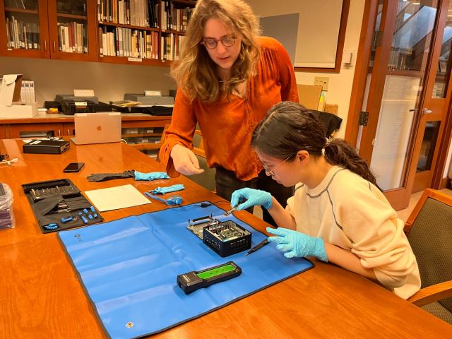 Two people work at a table disassembling an electronic device, one standing and pointing, the other seated and wearing blue gloves. Tools and equipment are spread out on the table.