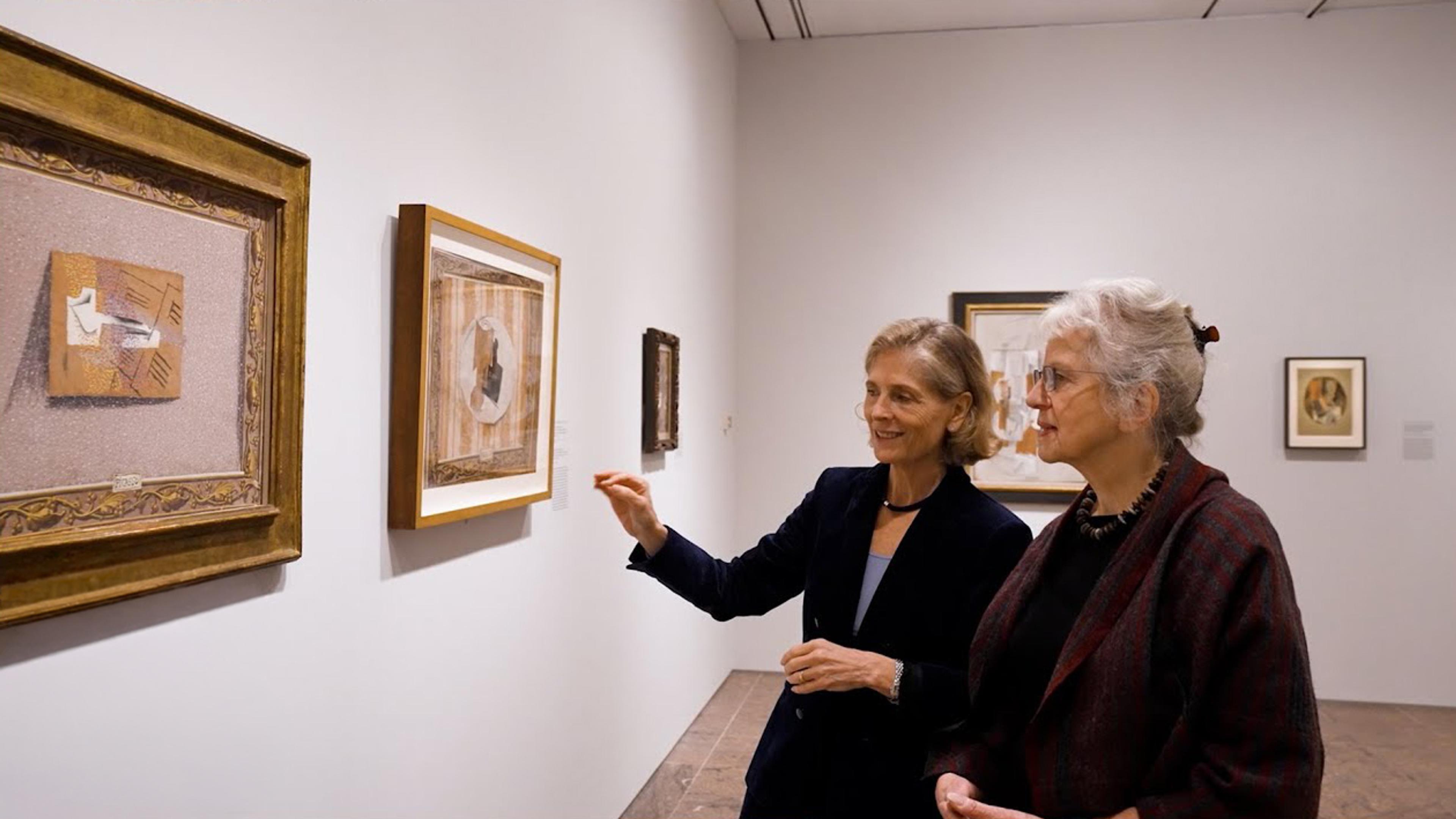Two women stand in a gallery looking at and discussing a painting on the wall in front of them.