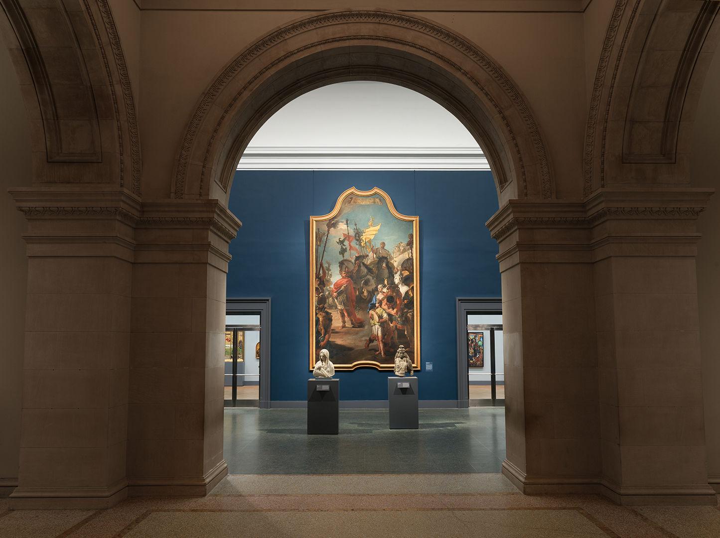 In-gallery view of Tiepolo's The Triumph of Marius. The imposing painting, set against a dark blue wall, is framed by a curved archway.