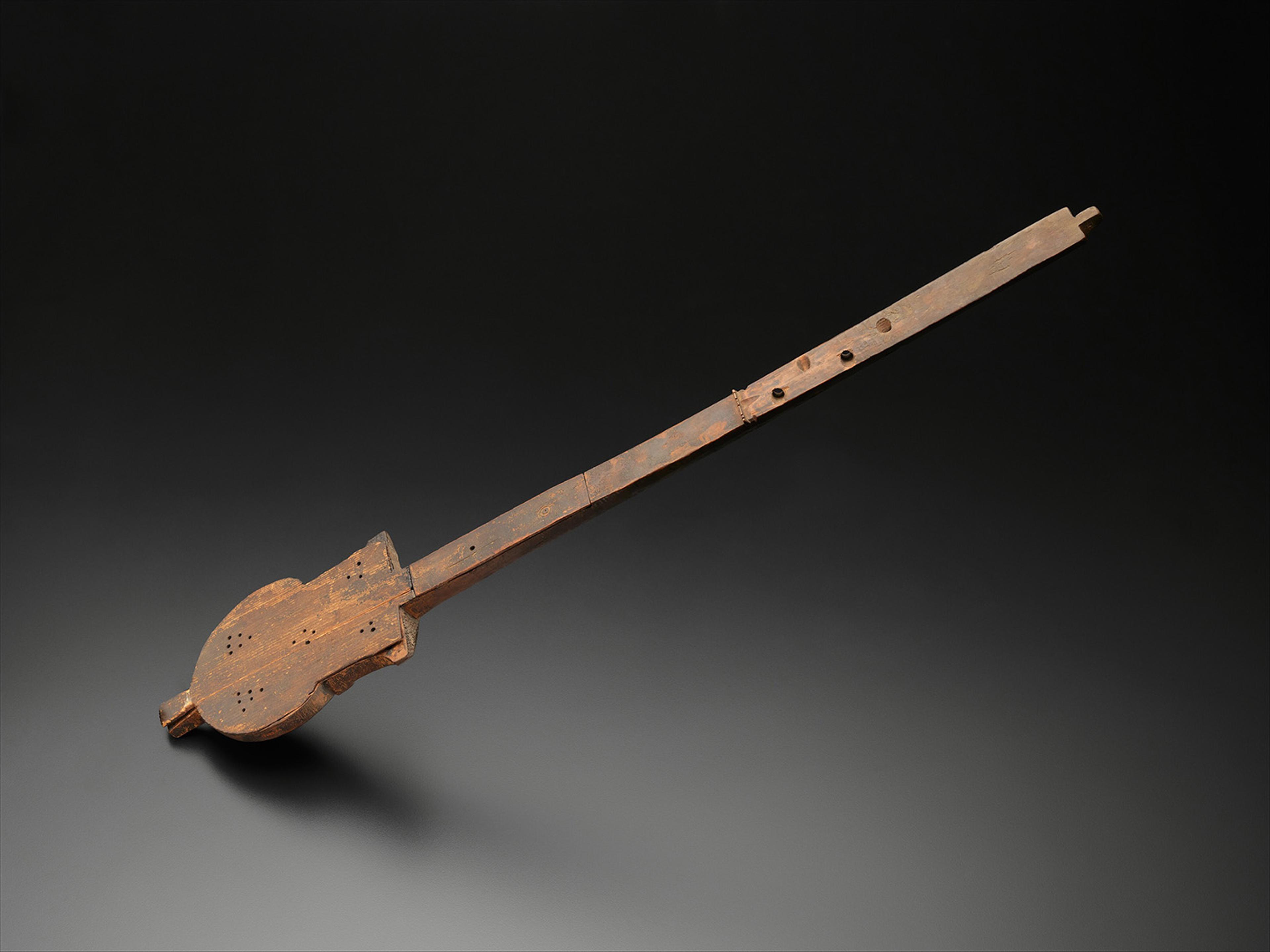 Old, rustic stringless wooden instrument on a dark background. It has a long, straight neck and a rounded, carved body, evoking a historical feel.