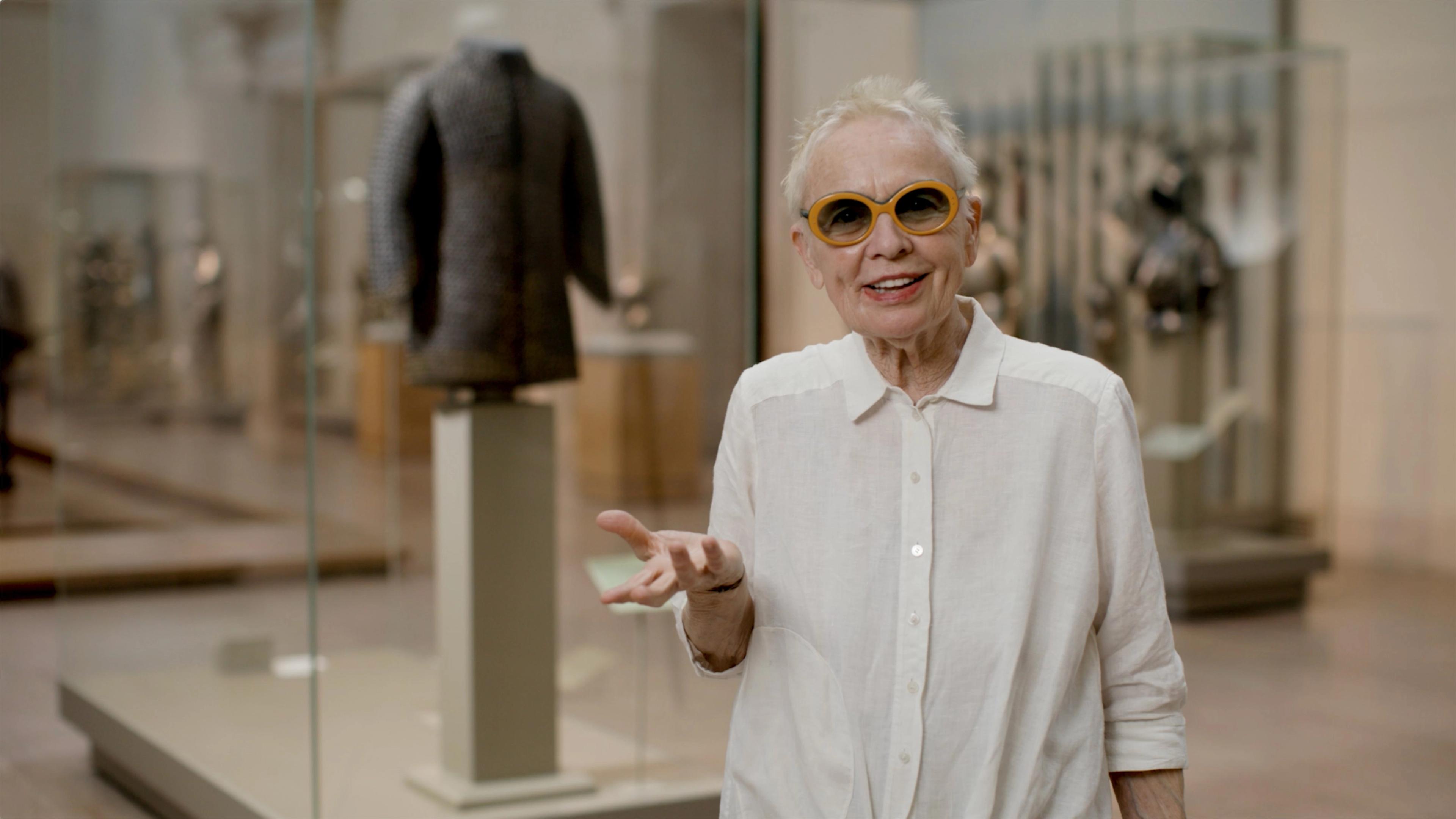An older, smiling woman gestures in front of herself. In the background is a blurred Arms and Armor gallery.