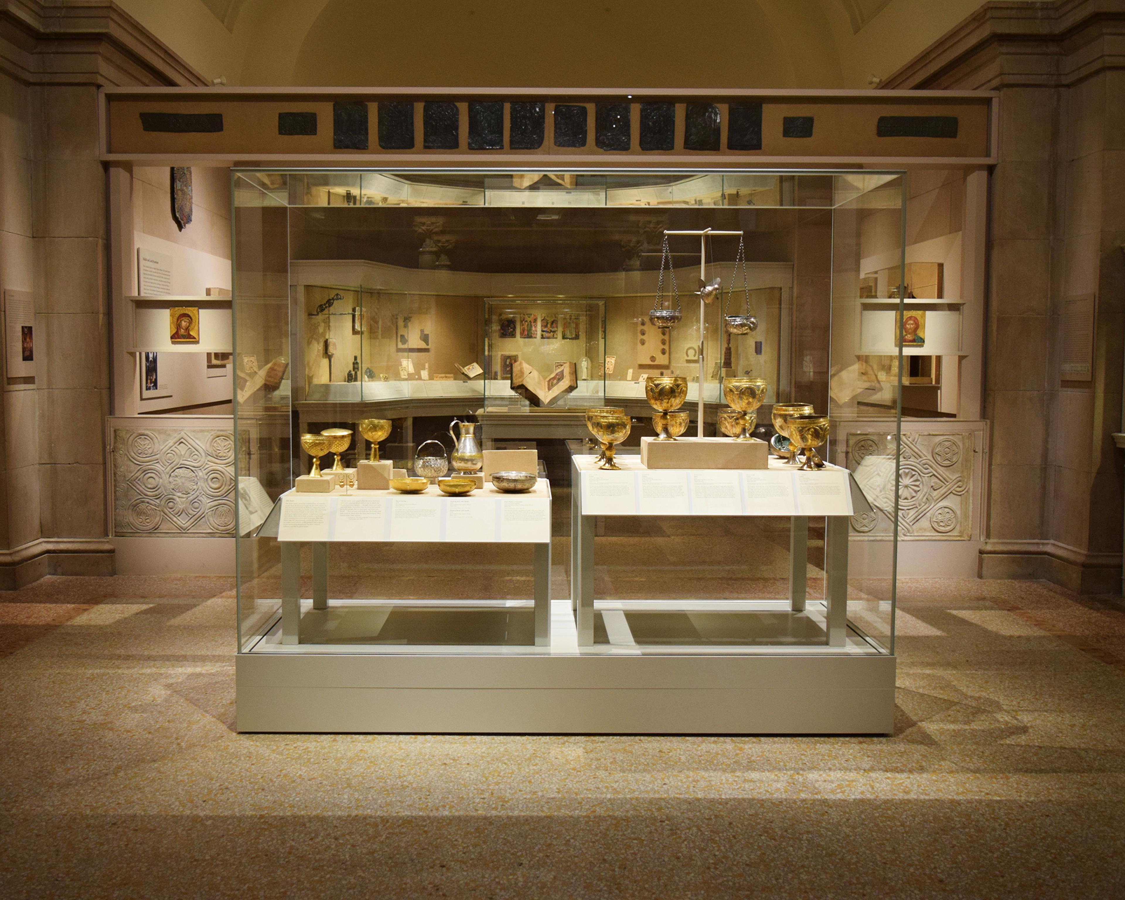A display case in a large gallery holds several gold cups, bowls, and plates