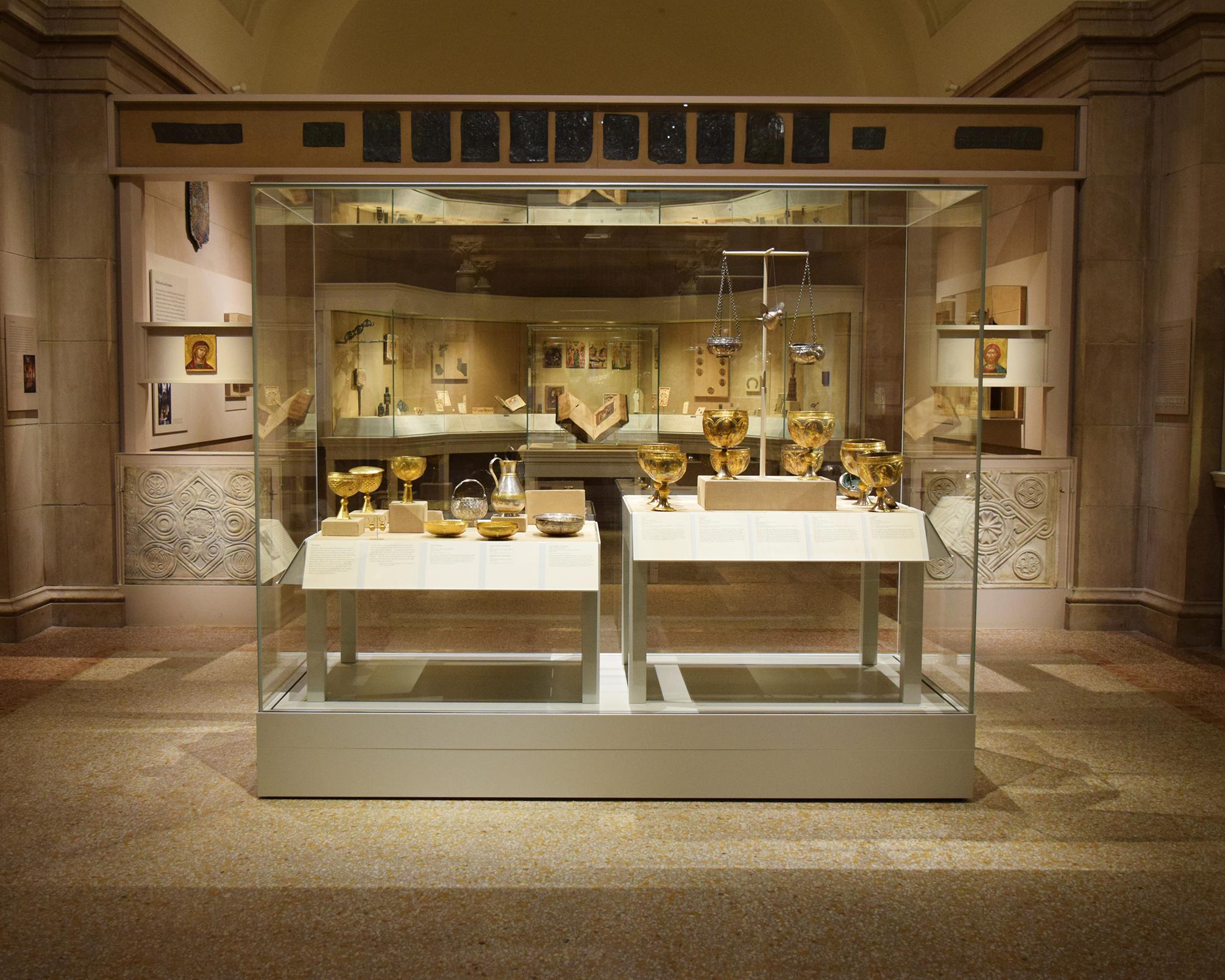 A display case in a large gallery holds several gold cups, bowls, and plates