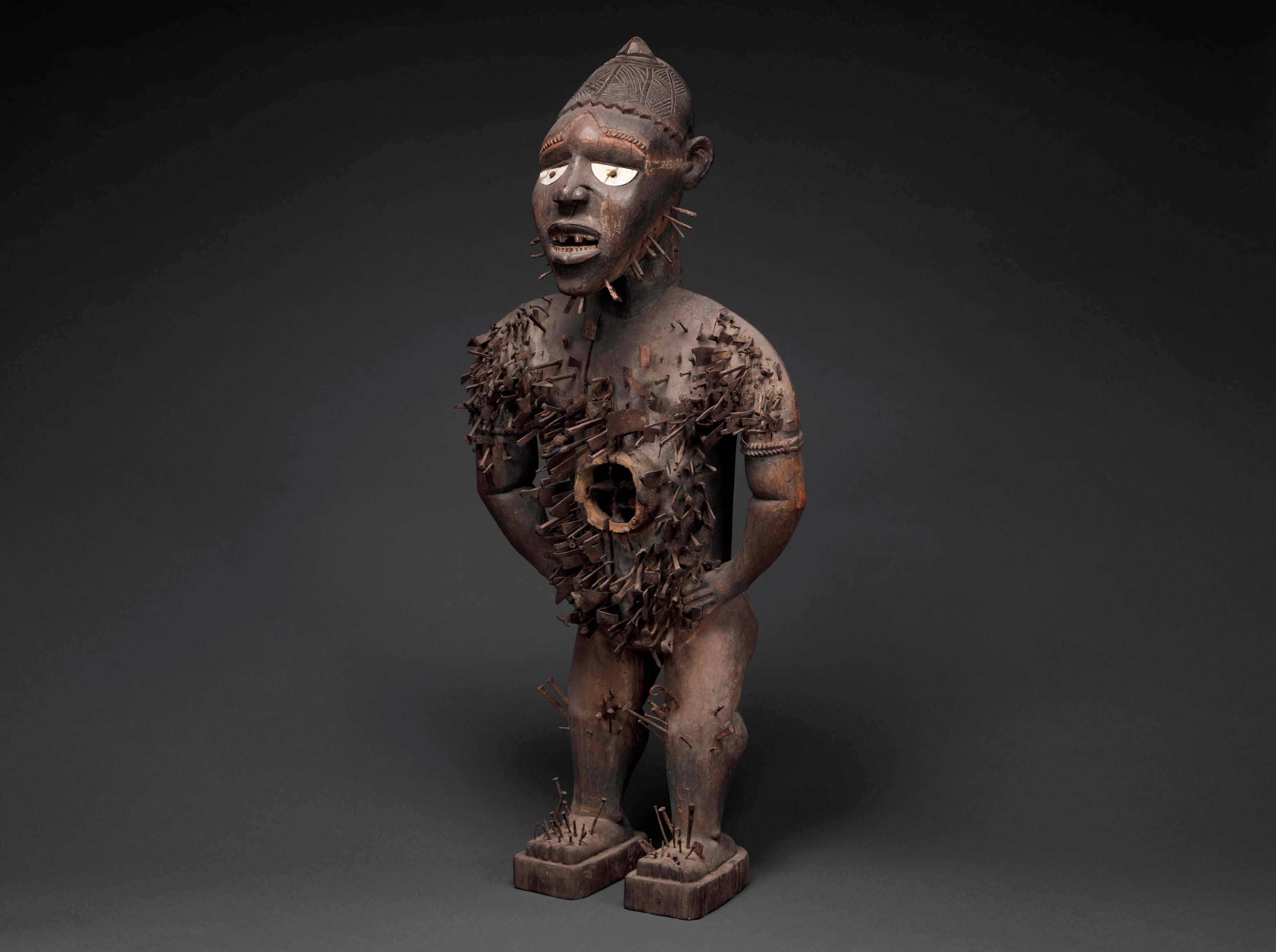 Carved wooden figure with inlaid eyes and a textured surface studded with nails.