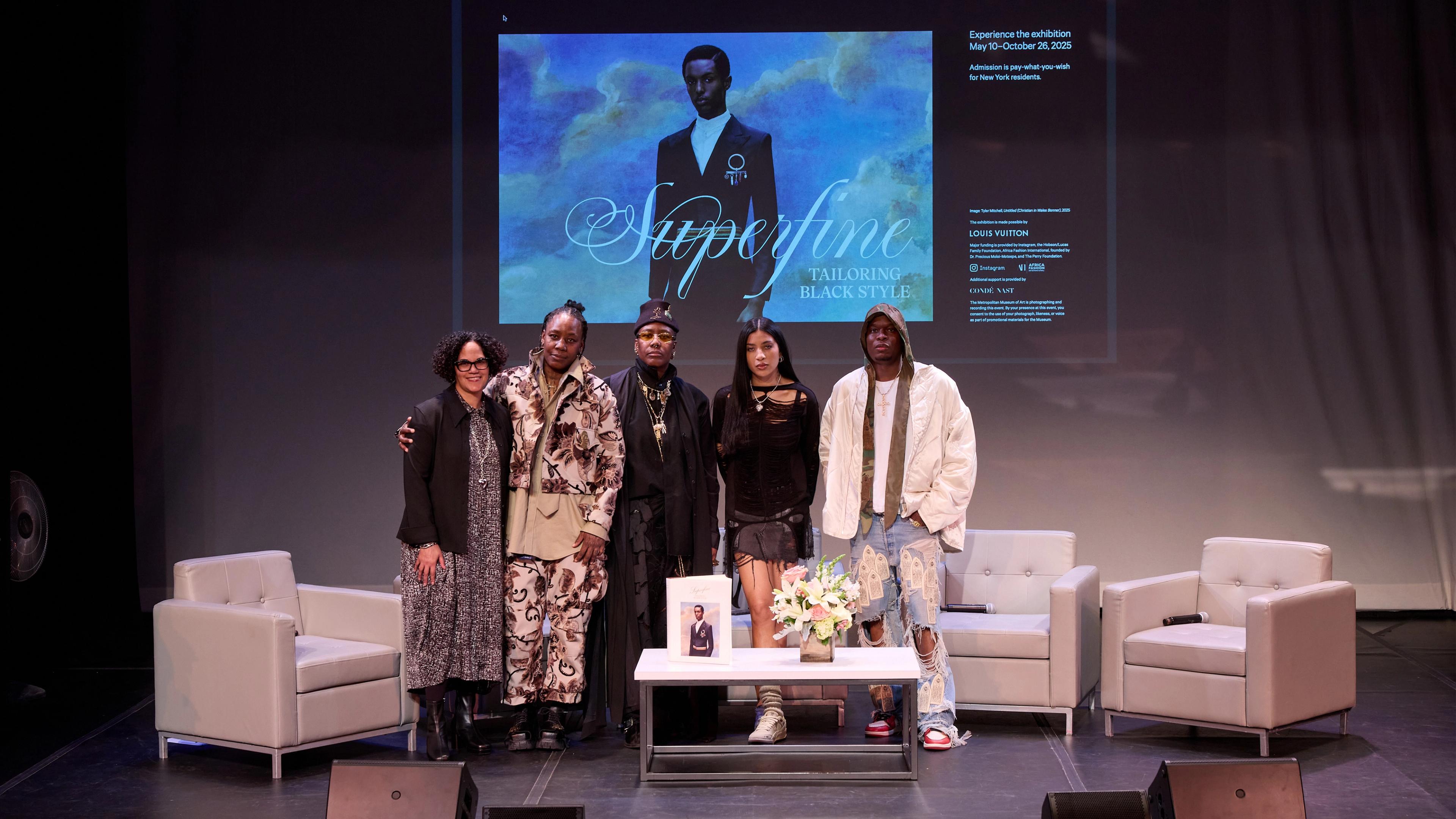 Five people stand on a stage, smiling warmly. Behind them is a large screen displaying "Superfine: Tailoring Black Style." The atmosphere is welcoming.