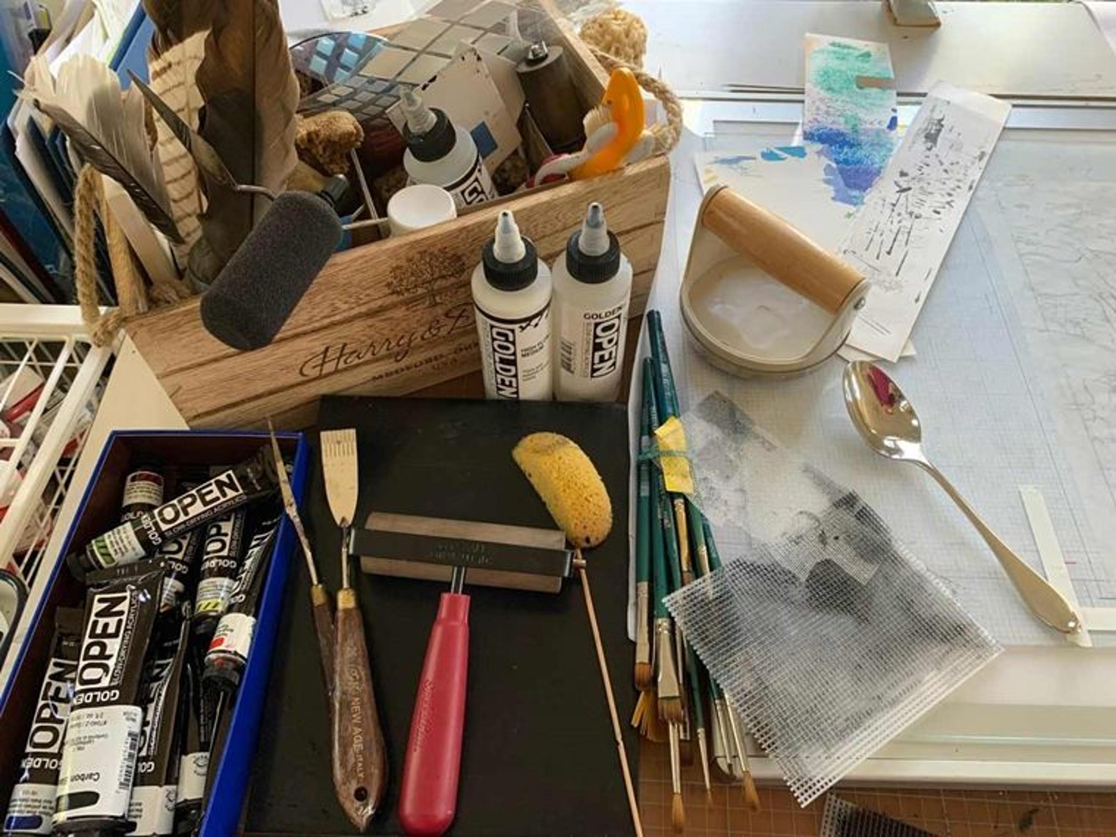 A collection of art supplies, including inks, paints, sponges, knives, and a palm-sized disc-shaped object with a wooden handle.
