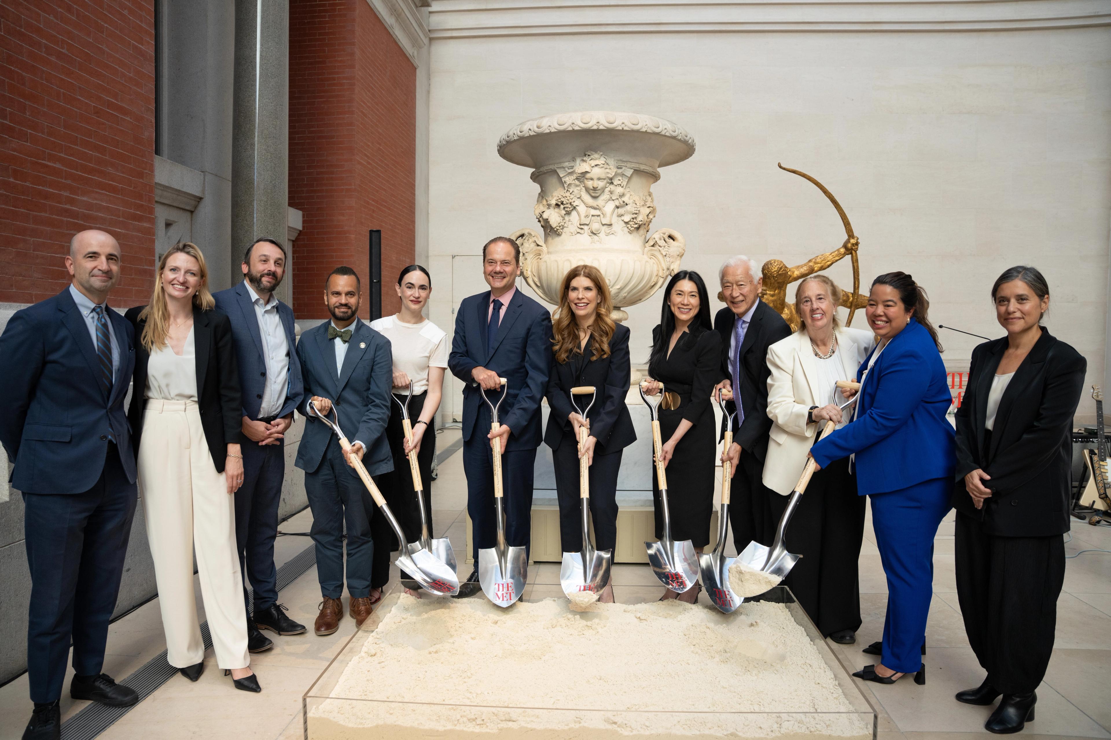 A group of twelve people in business attire pose with ceremonial "THE MET" shovels, digging into a sand-filled display case for a museum groundbreaking, with a marble urn and bronze archer statue behind them.