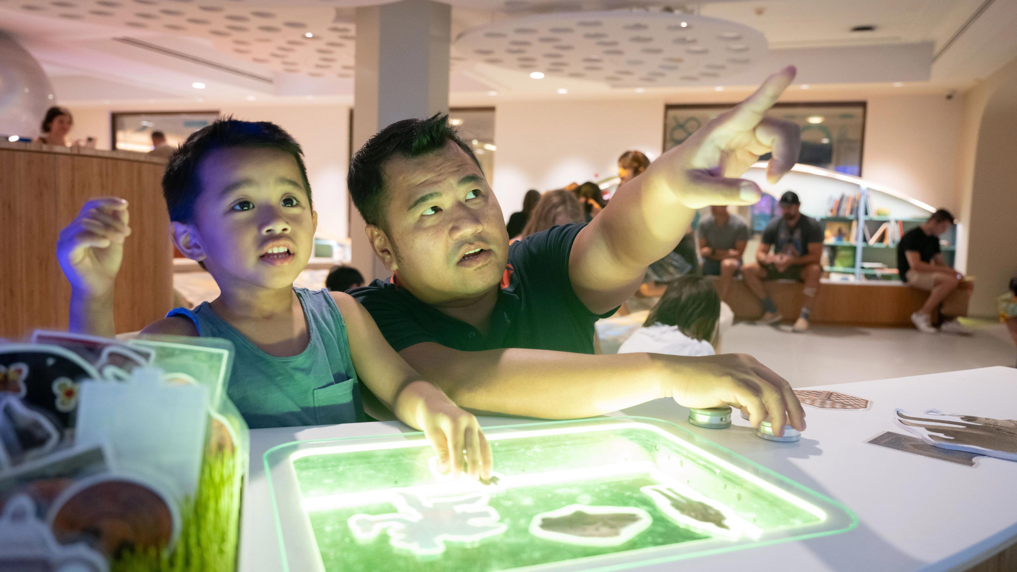 A father and son sit at a playful interactive table with green light, father is pointing up.