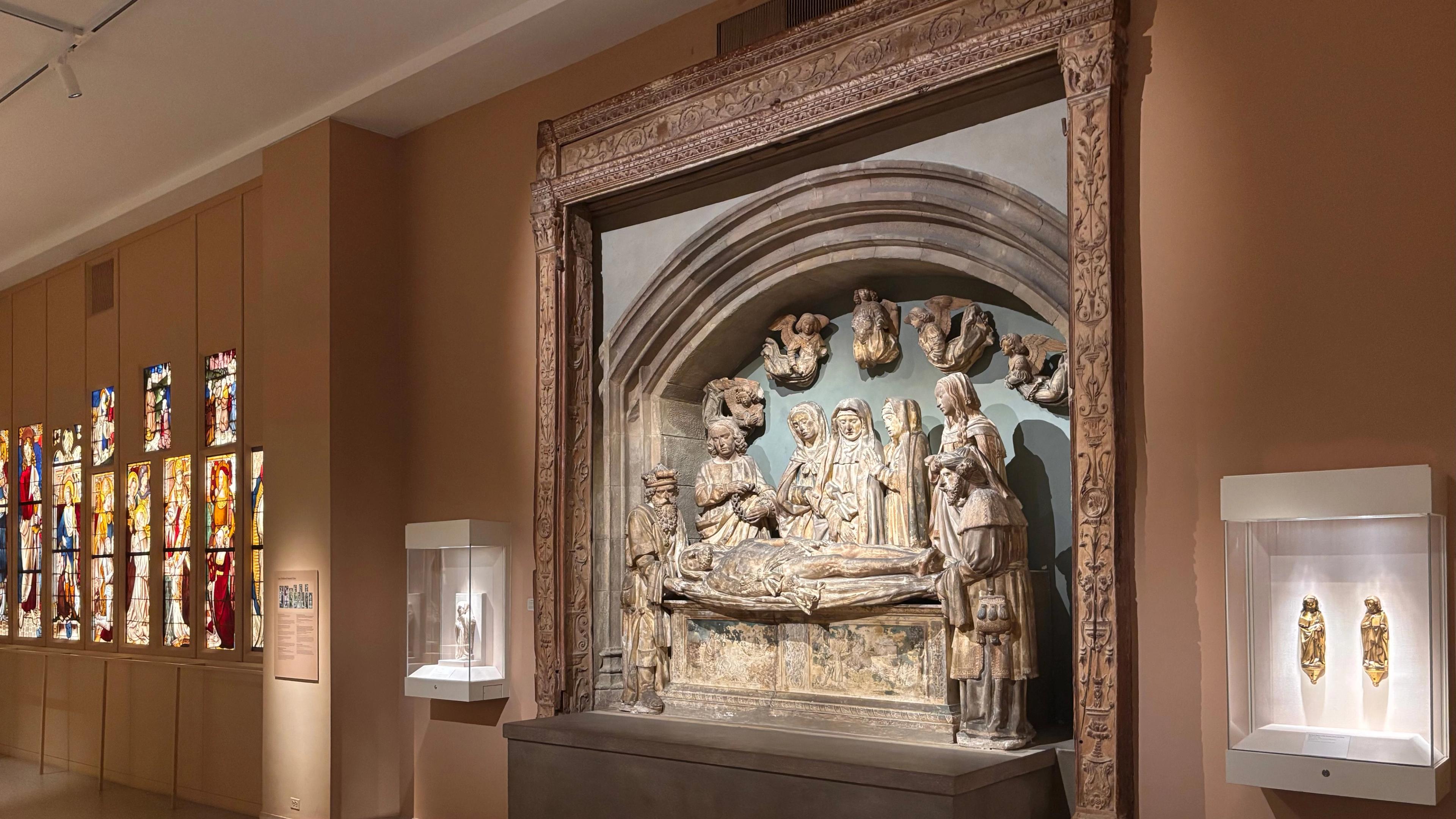 Gallery view of colorful stained glass windows, two vitrines with gold figures and a central, large-scale stone sculpture.