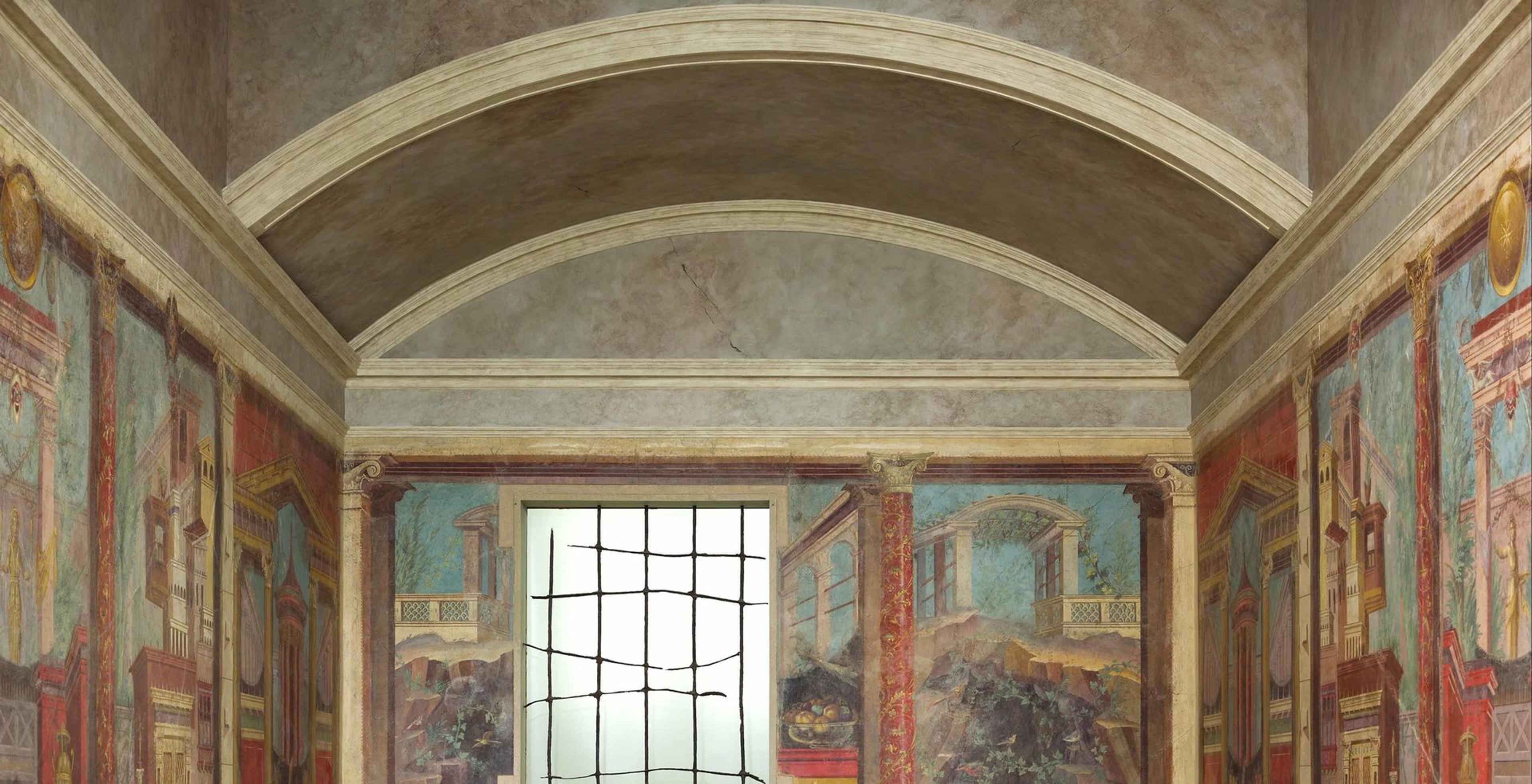 An ornate Roman-style room with painted frescoes depicting architectural and natural scenes, framed by columns and arches, with a central window featuring a grid design letting in soft light.
