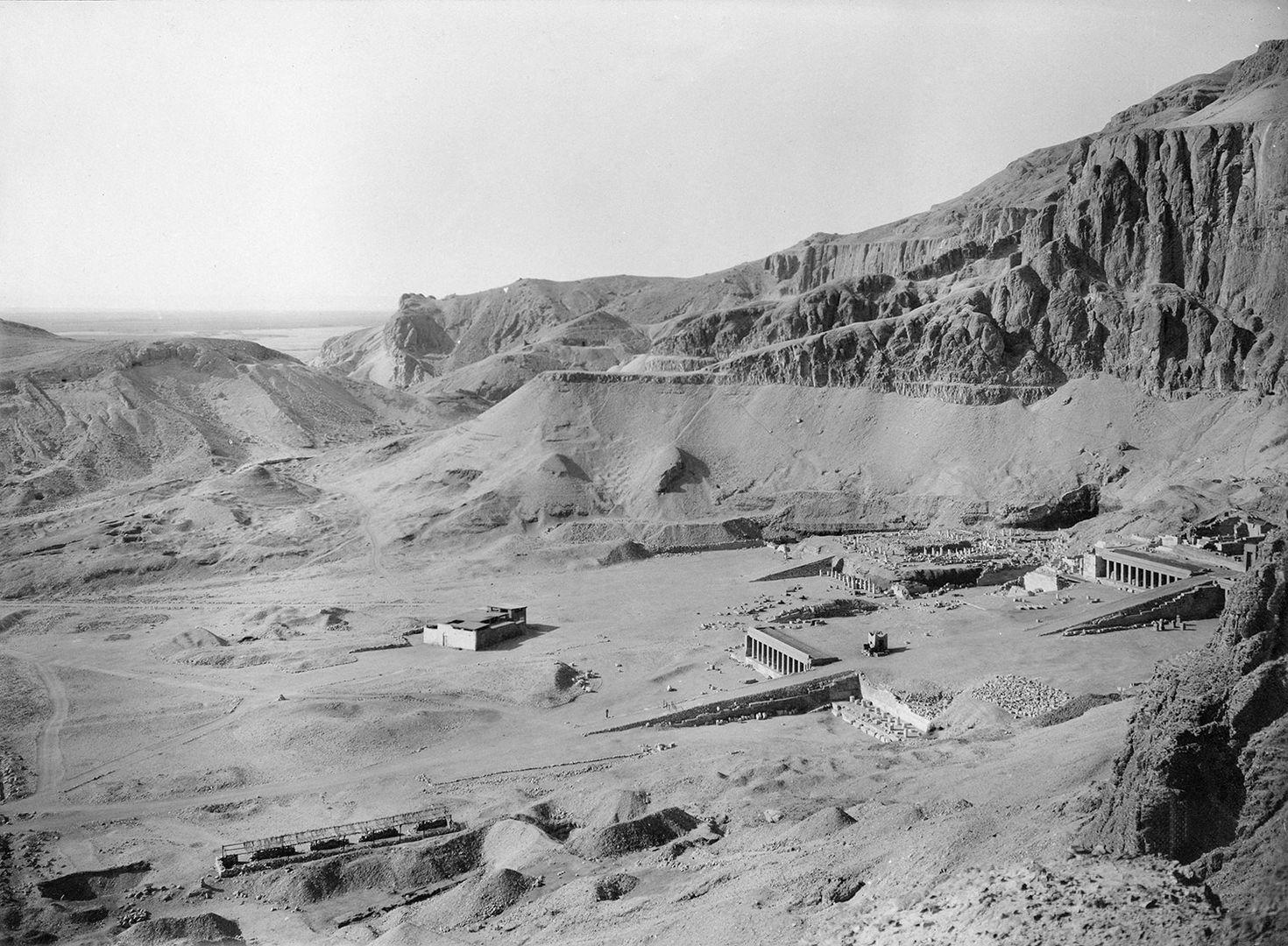A view from high up on a cliff looking down over a desert valley in which the ruins of a large temple can be seen.