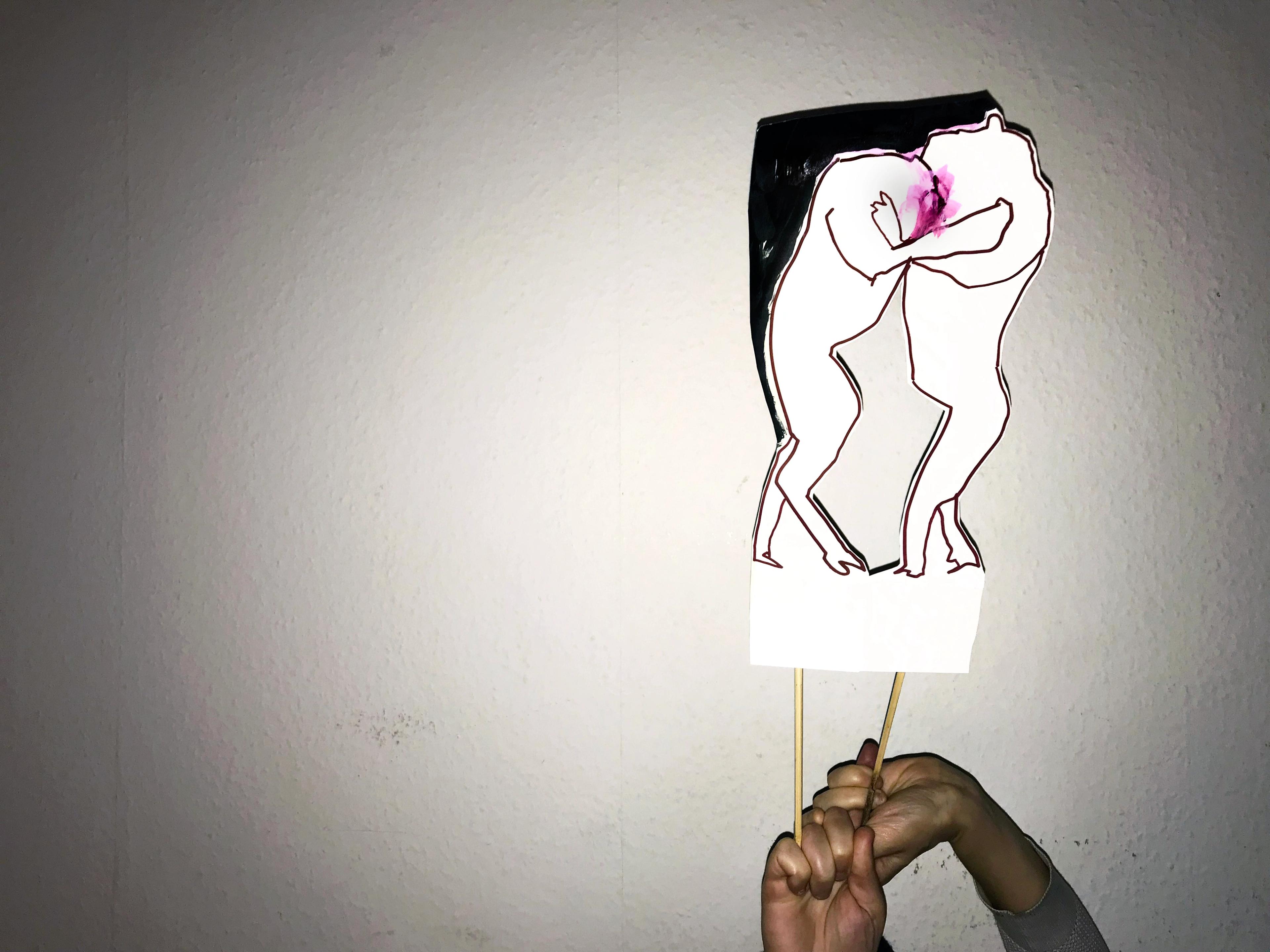 Two hands holding sticks on which are attached a cut out of two white lion figures embracing with a pink smudge between their faces