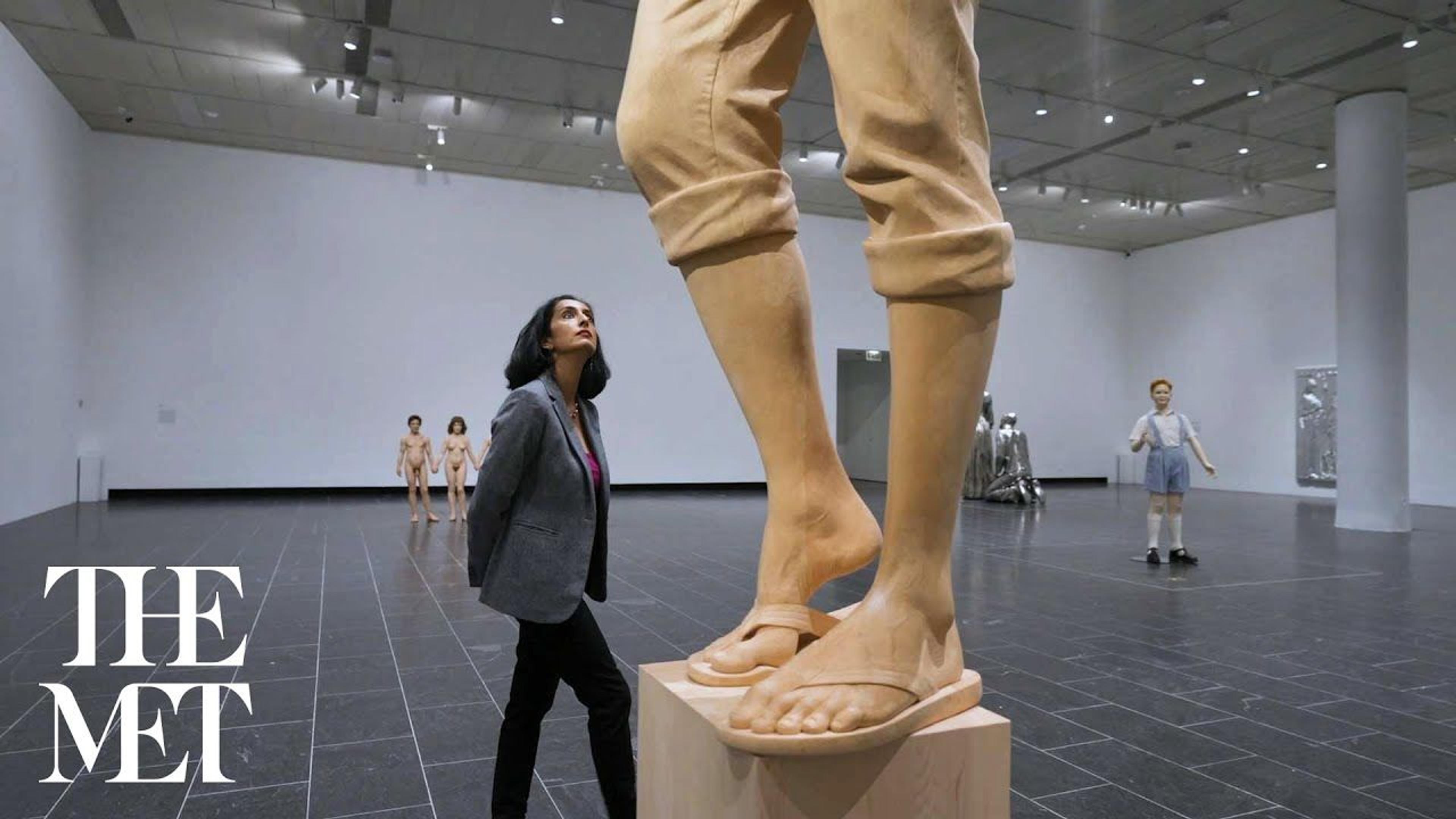 A woman walks through an exhibition space, looking up at an oversize sculpture of a man in rolled-up jeans, carved from cypress wood.