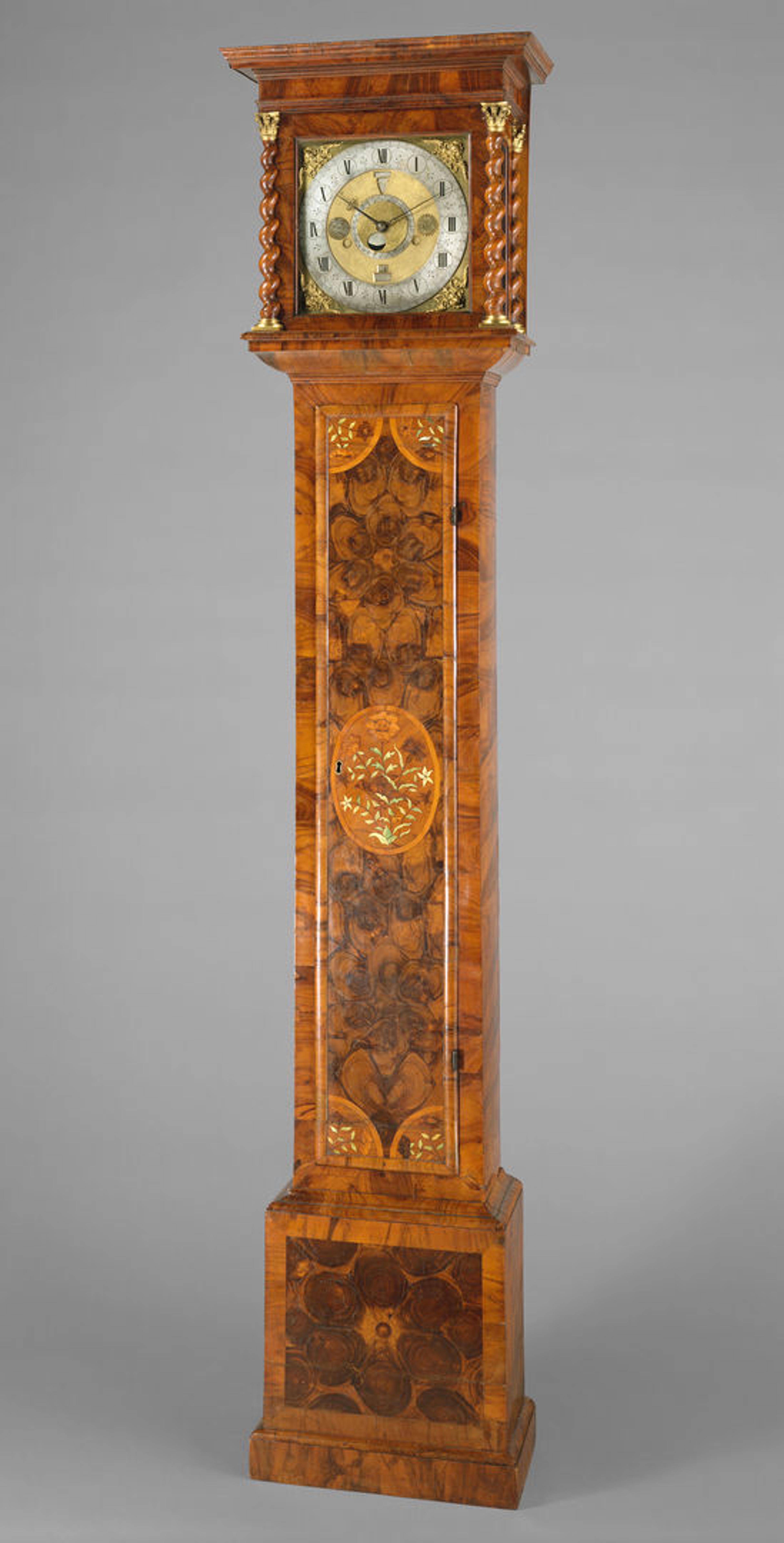 Longcase clock. The shading of light and dark browns in the wood creates a marbled effect, and gold details are inlaid around the clock face and throughout the paneling.