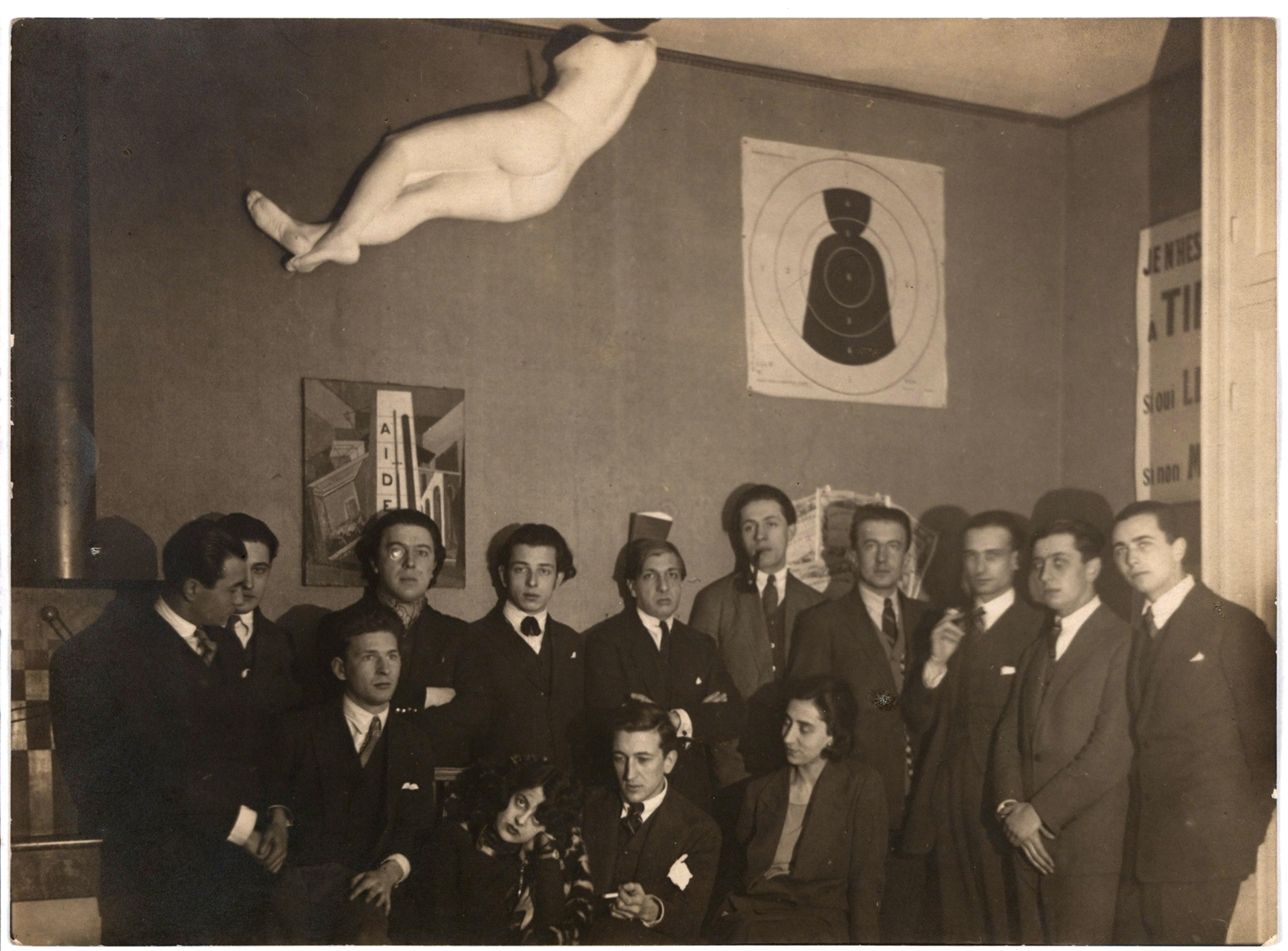 Black-and-white portrait of a group of people wearing dark suits with a nude mannequin attached to the wall above them.