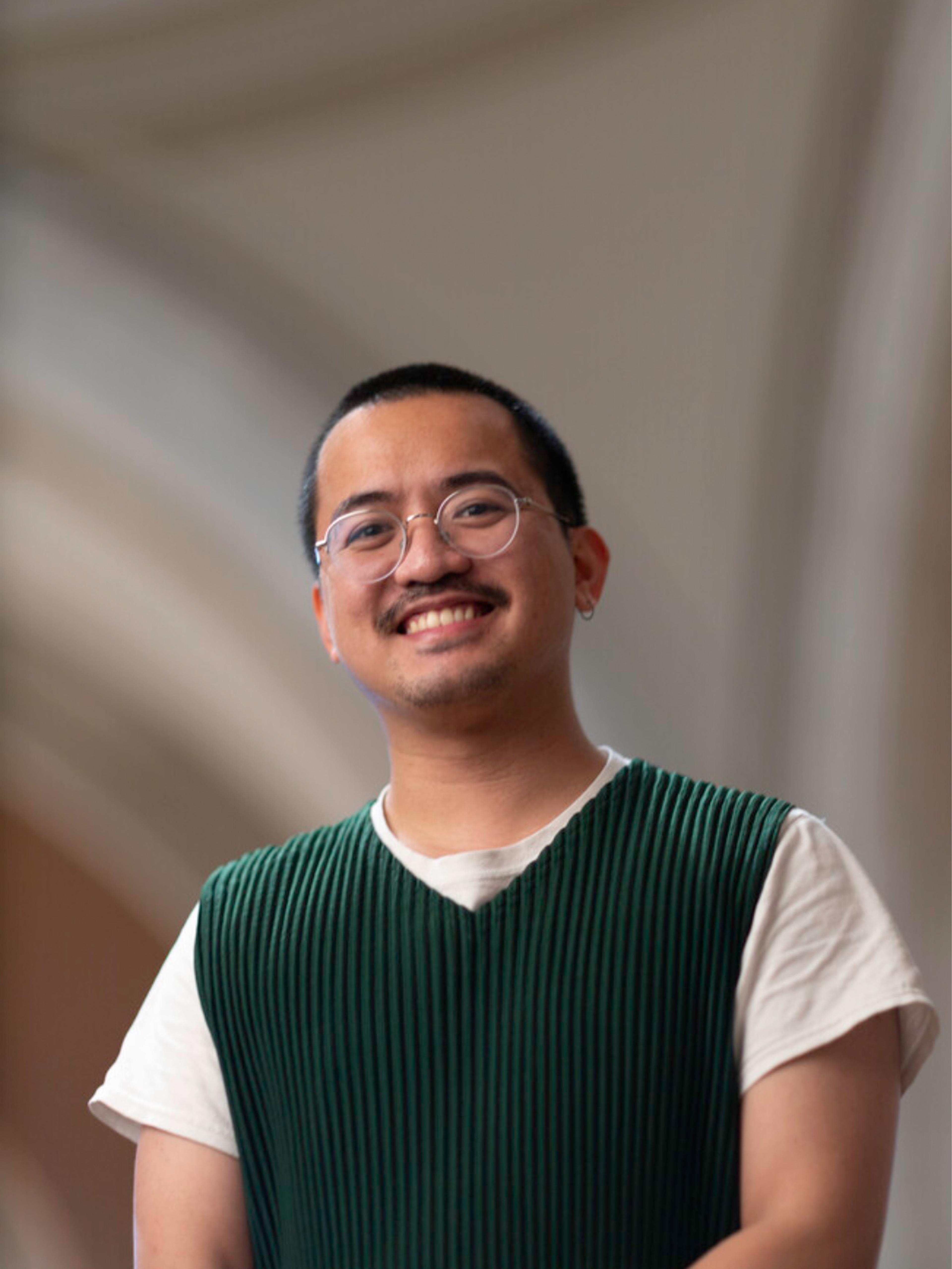 A smiling man in round glasses and a dark green ribbed sweater vest over a white T-shirt stands against a softly lit, neutral background.