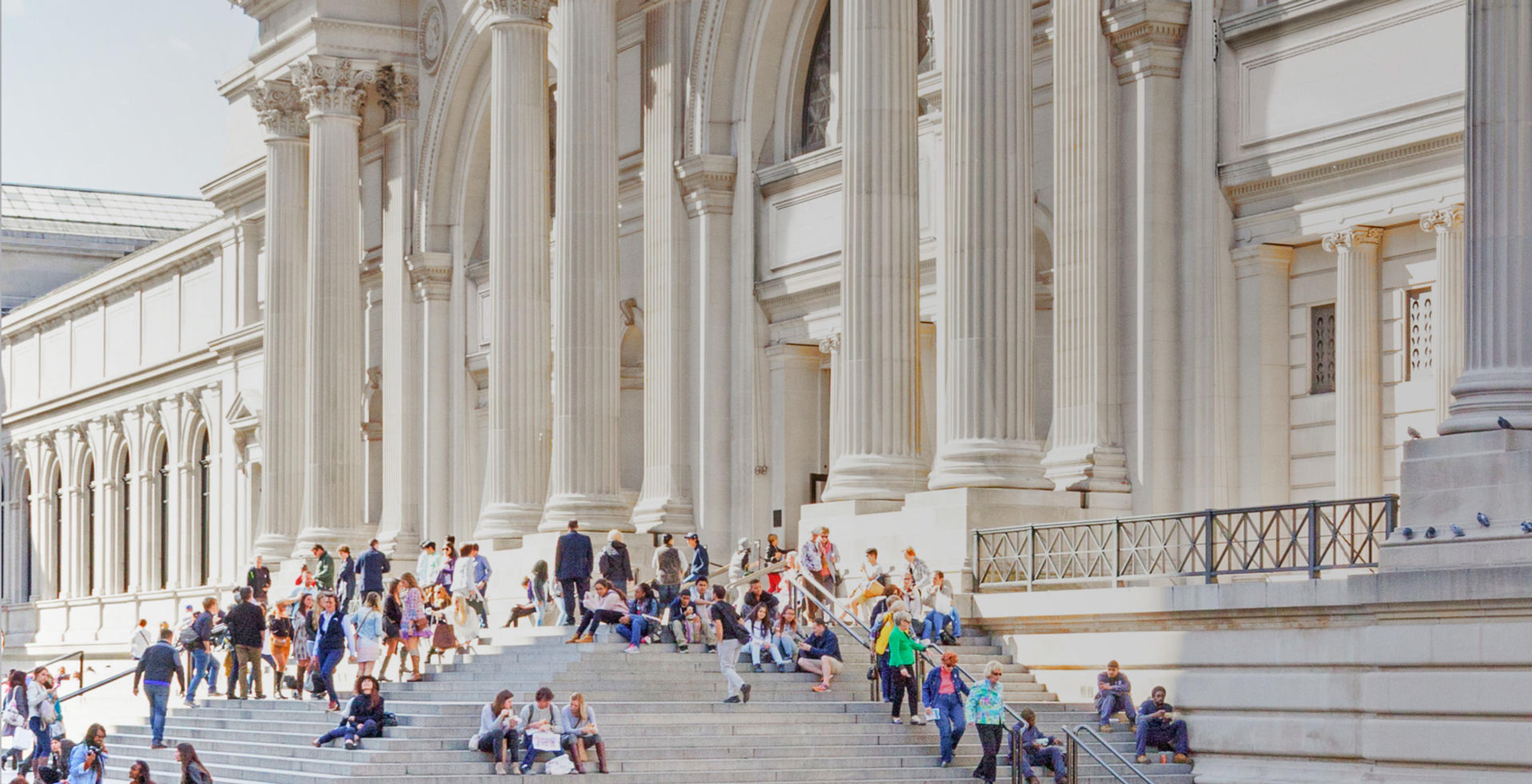 South-facing view of The Met exterior steps from the 5th Ave plaza. Sunlight hits the building facade, and the steps bustle with seated and standing visitors.