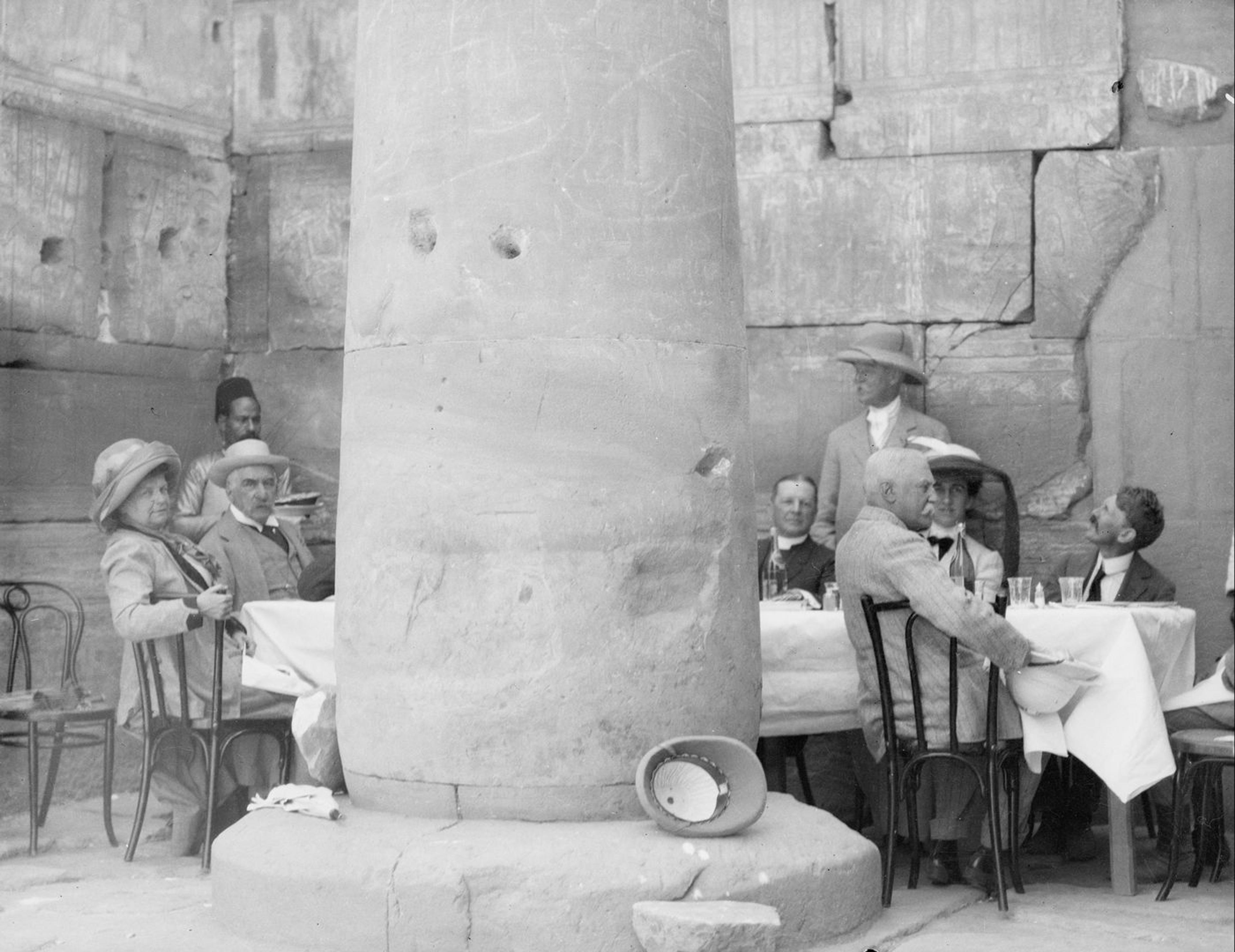 Old photograph of men and women dining at a table near a stone excavation site