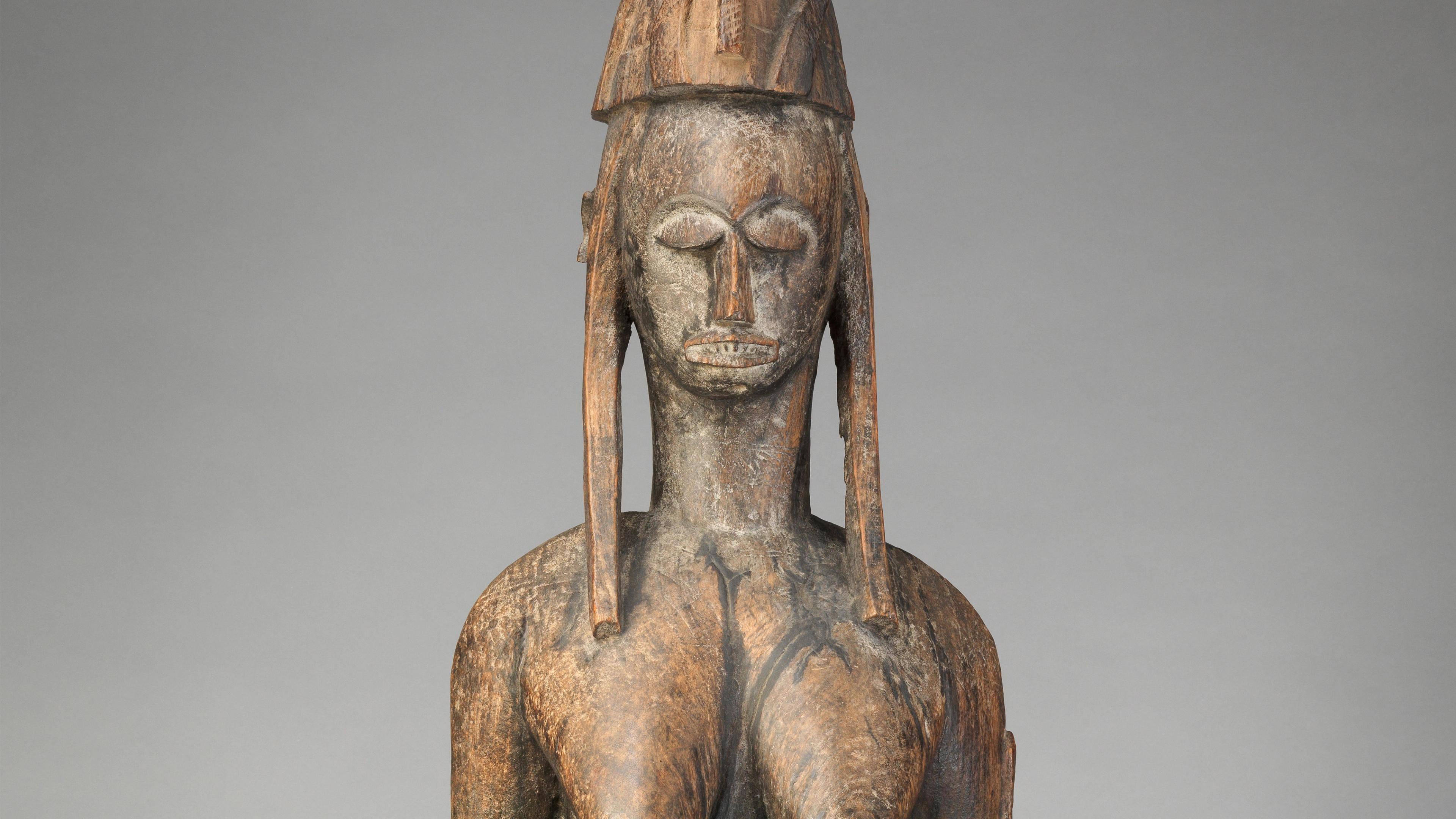 Wooden sculpture of a woman wearing an amulet-laden hat