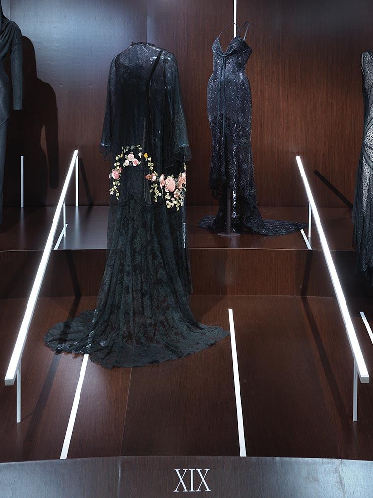 Installation view of the Versace dress displayed on an invisible mount in <em>About Time: Fashion and Duration</em> next to a flowing, black dress with colorful embroidery detail around the hips and lower back