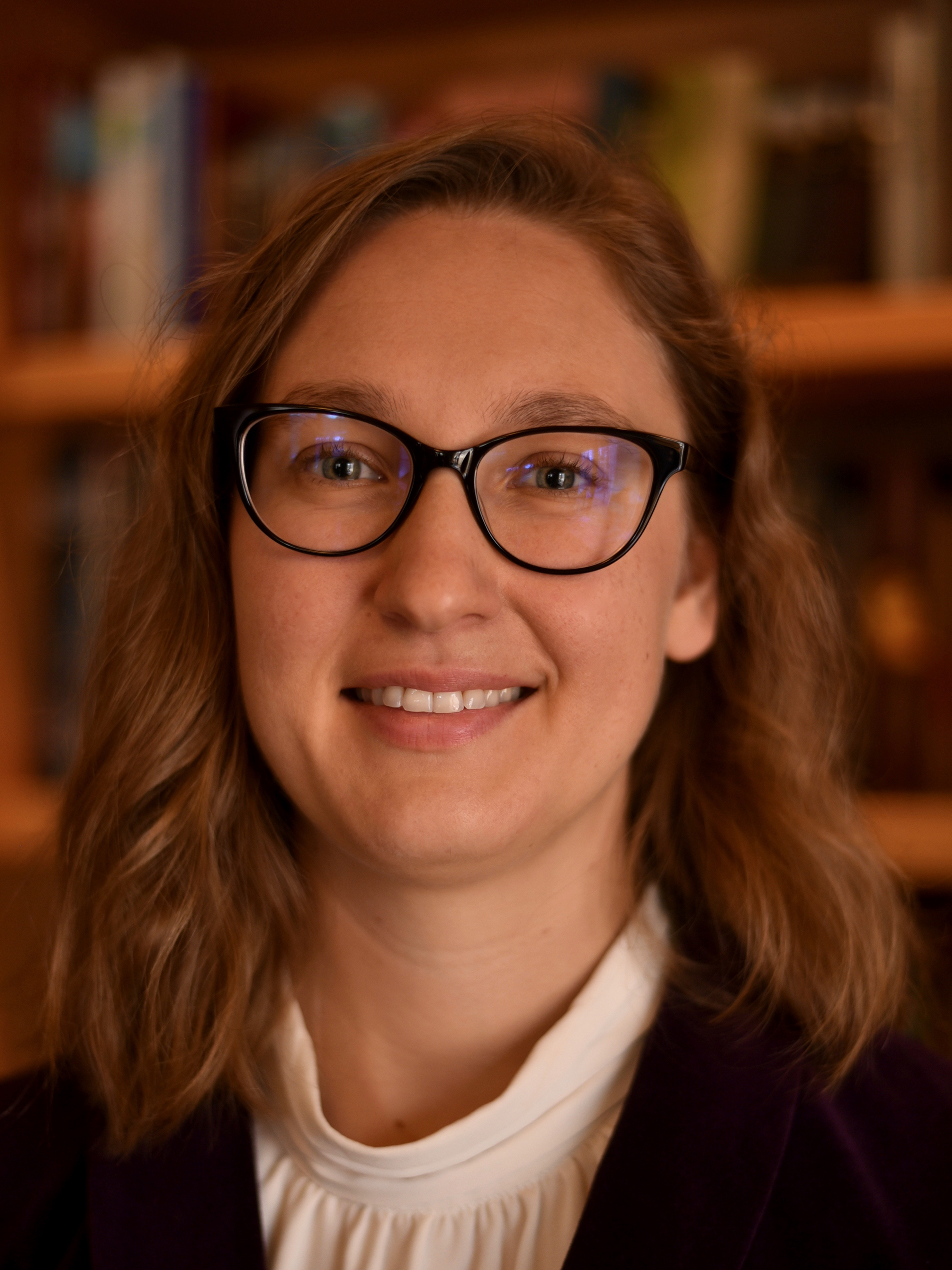 A woman wearing glasses and a blazer poses in front of a bookcase