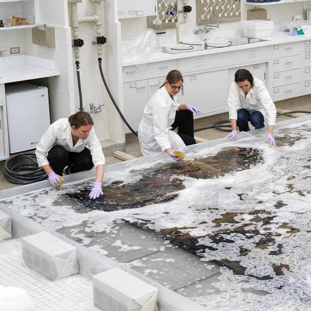 5 individuals in white lab coats kneel over a large, flat textile to clean it with foam and water, using sponges and other tools.