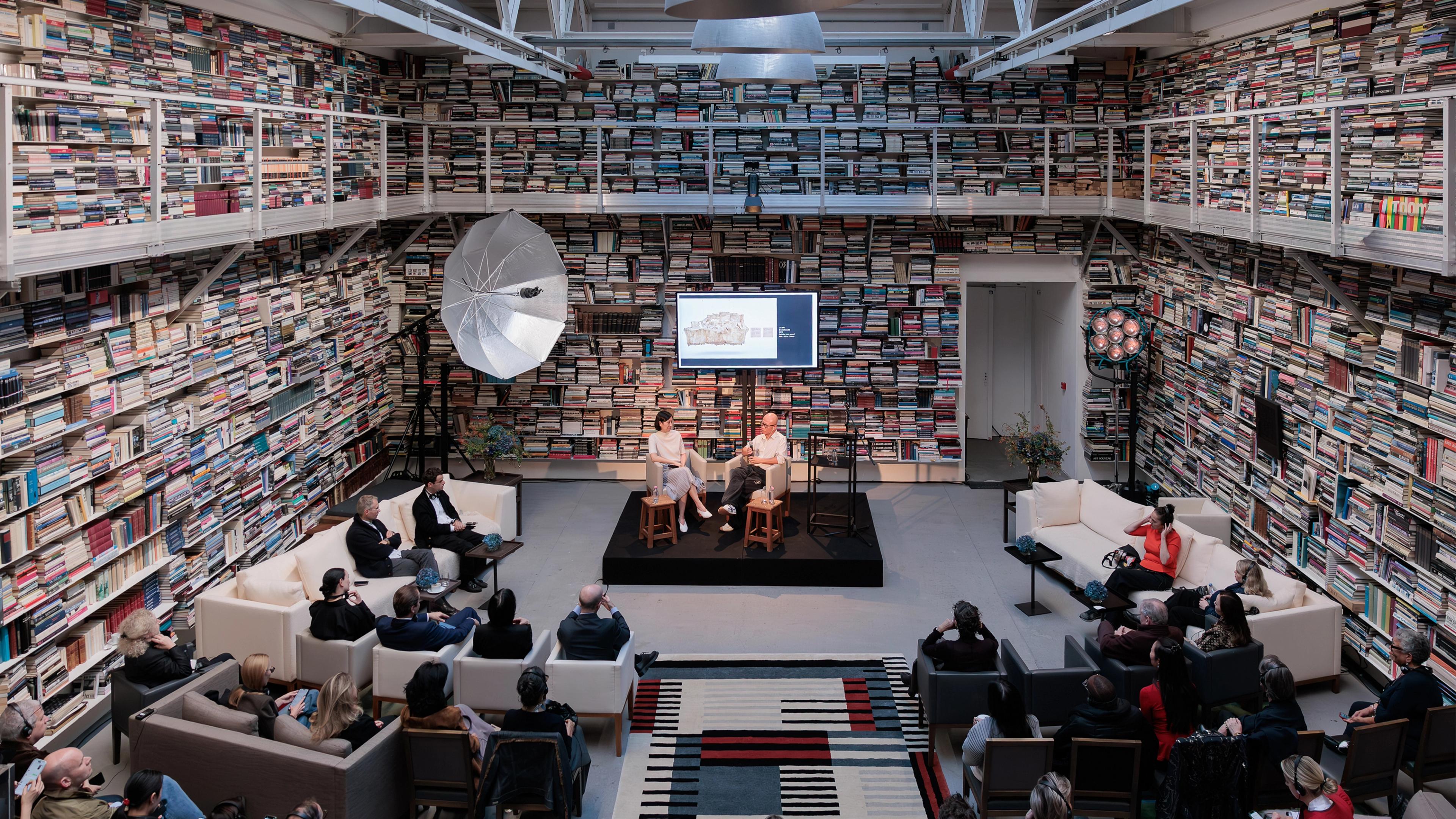 Two speakers seated on a small raised platform surrounded by books, while an audience listens attentively in a cozy setting.