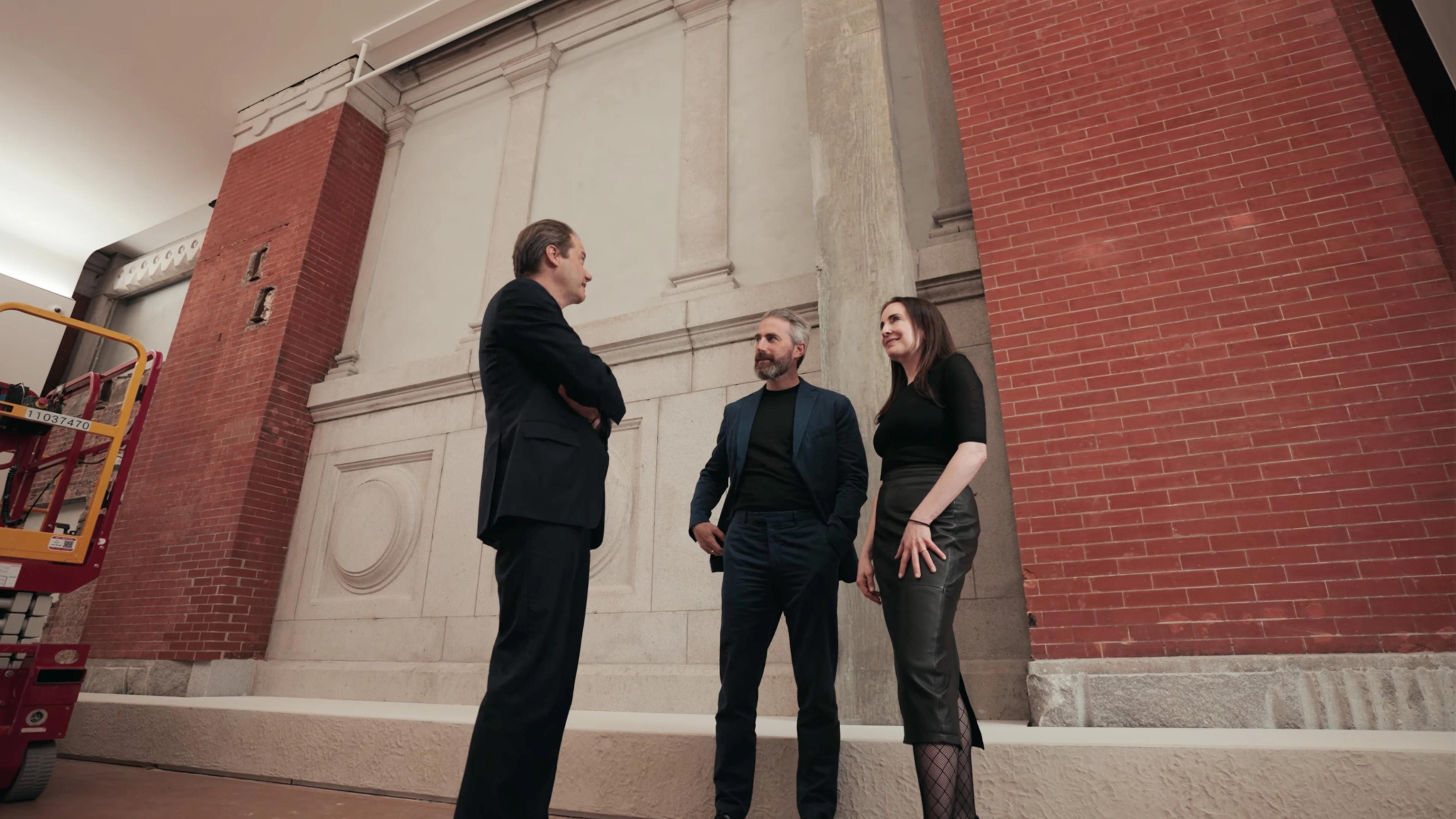 A woman and two men in formal attire stand conversing in front of a classical stone wall with columns