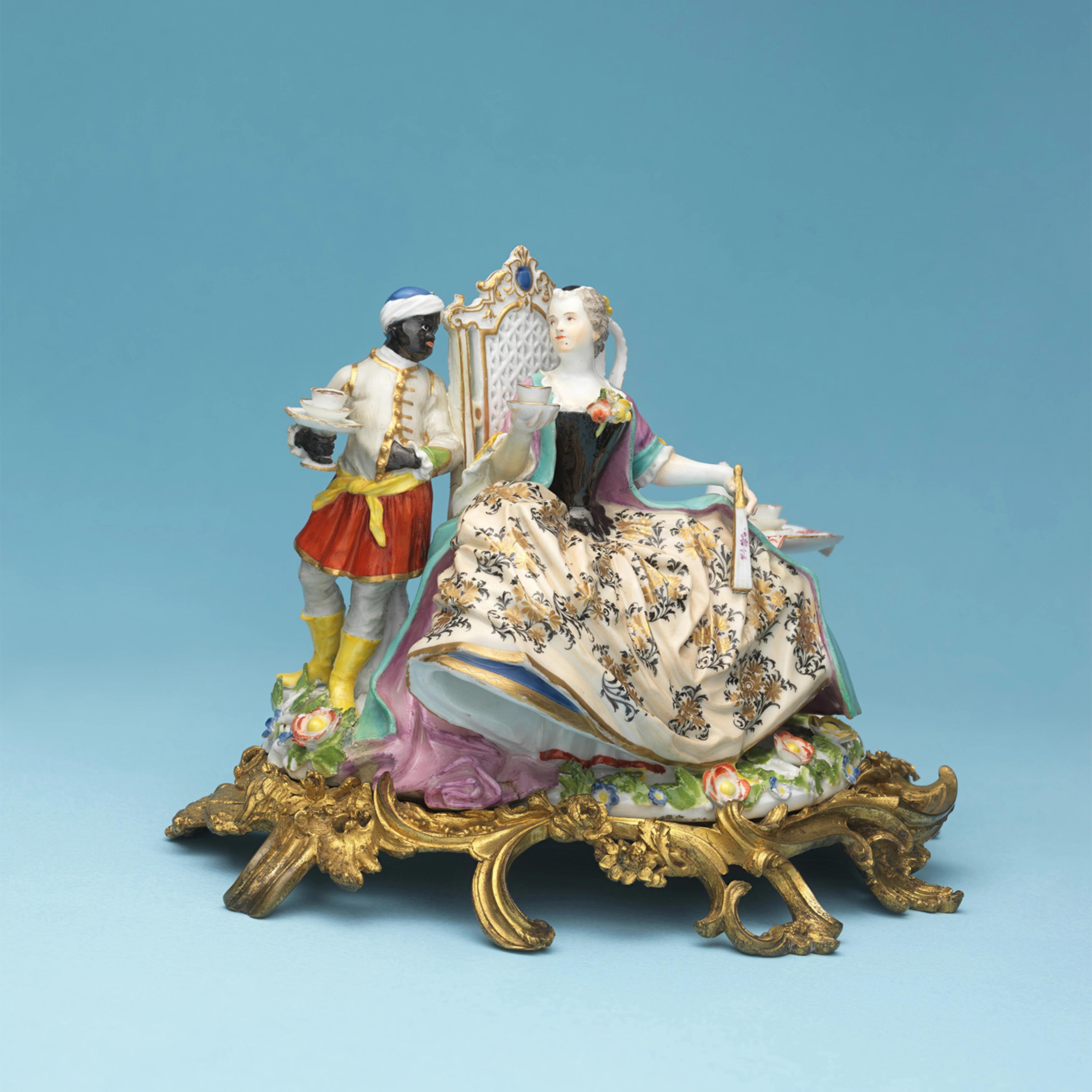 Porcelain figurine depicting an 18th-century scene with a servant holding a tray of cups and a seated aristocratic woman in floral attire, against a blue background.
