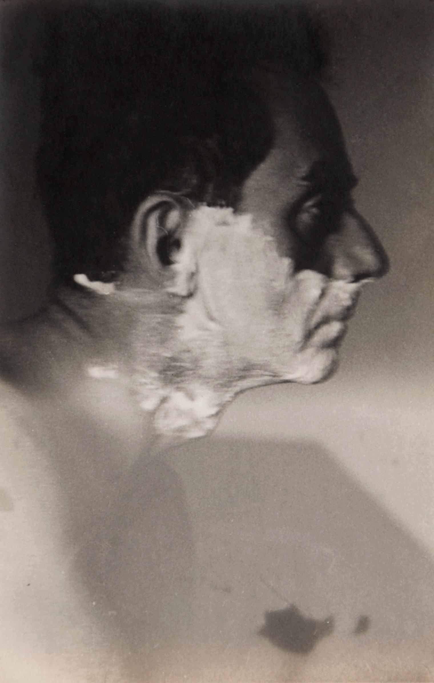 A grayscale photo of a man in profile, head turned left. His face and neck are partially covered in shaving cream. The expression appears contemplative.