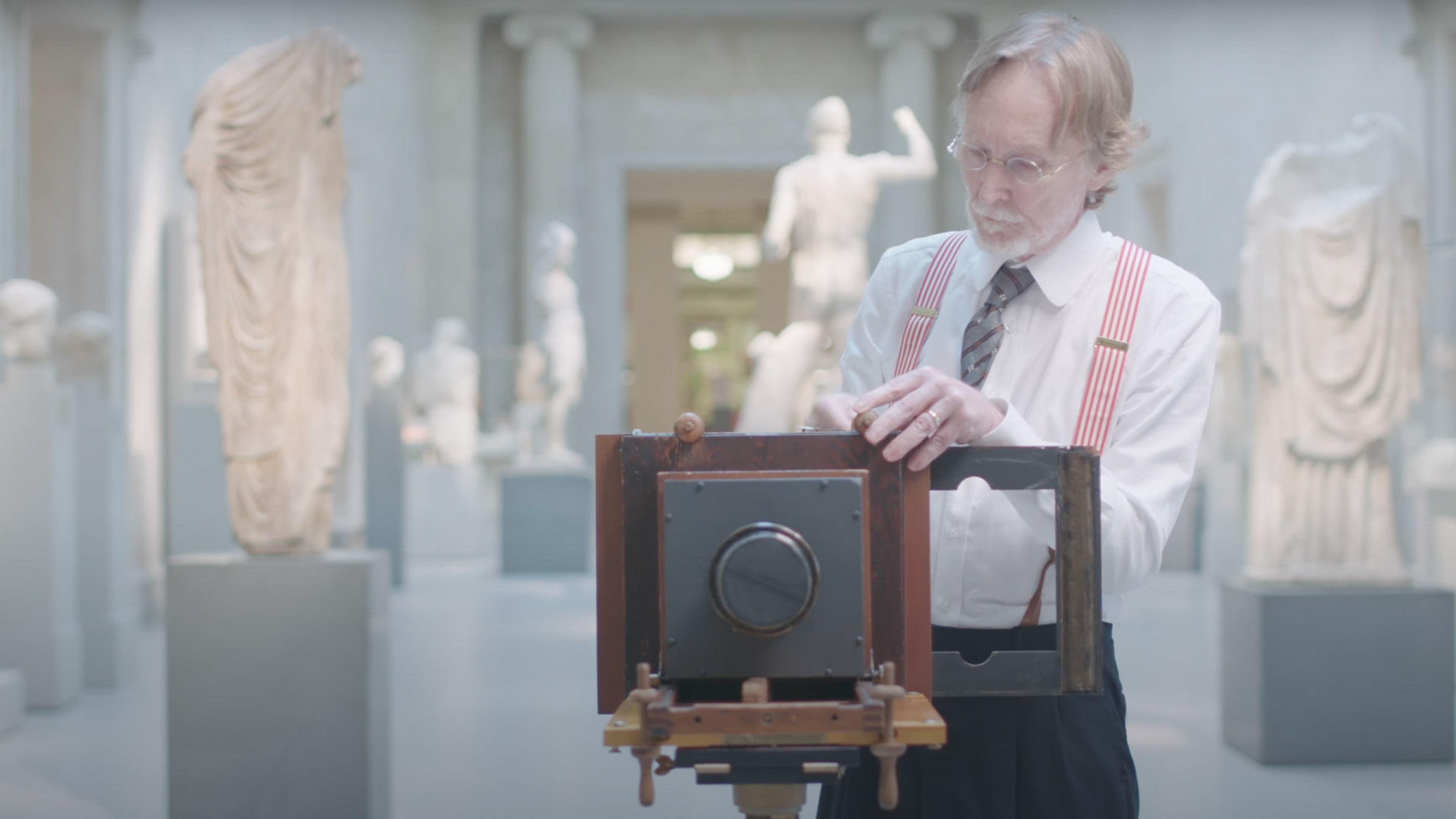 A man wearing blue pants, white shirt, striped tie and pink suspenders prepares a daguerreotype camera to take a photo.