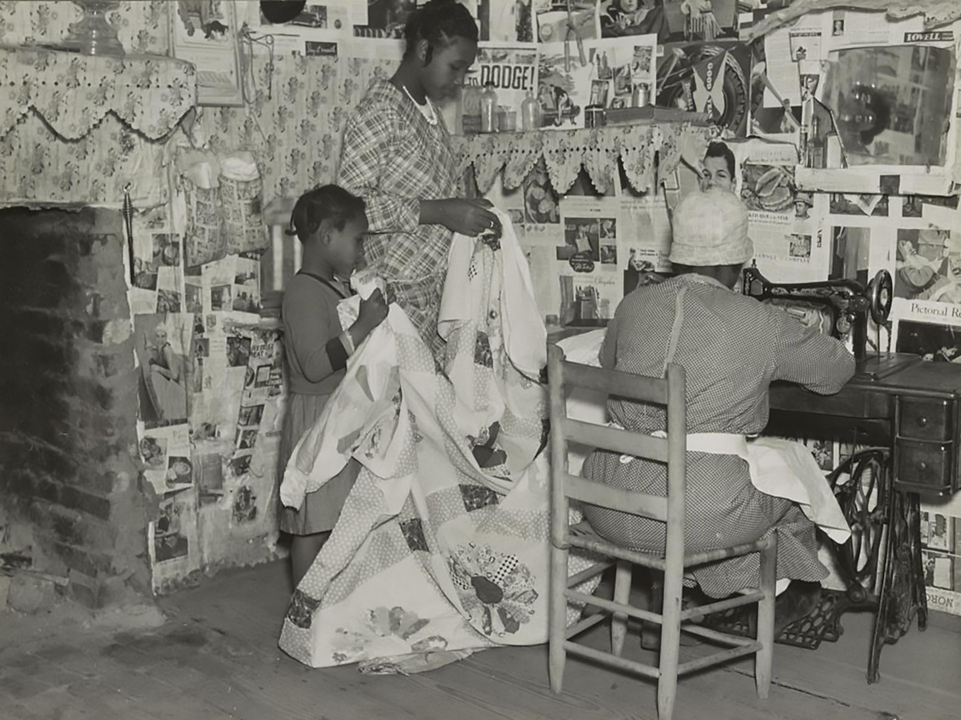 Black-and-white photograph of three people in a room decorated with wallpaper and advertisements. Two children hold up a white quilt being worked on by a figure at a sewing machine.