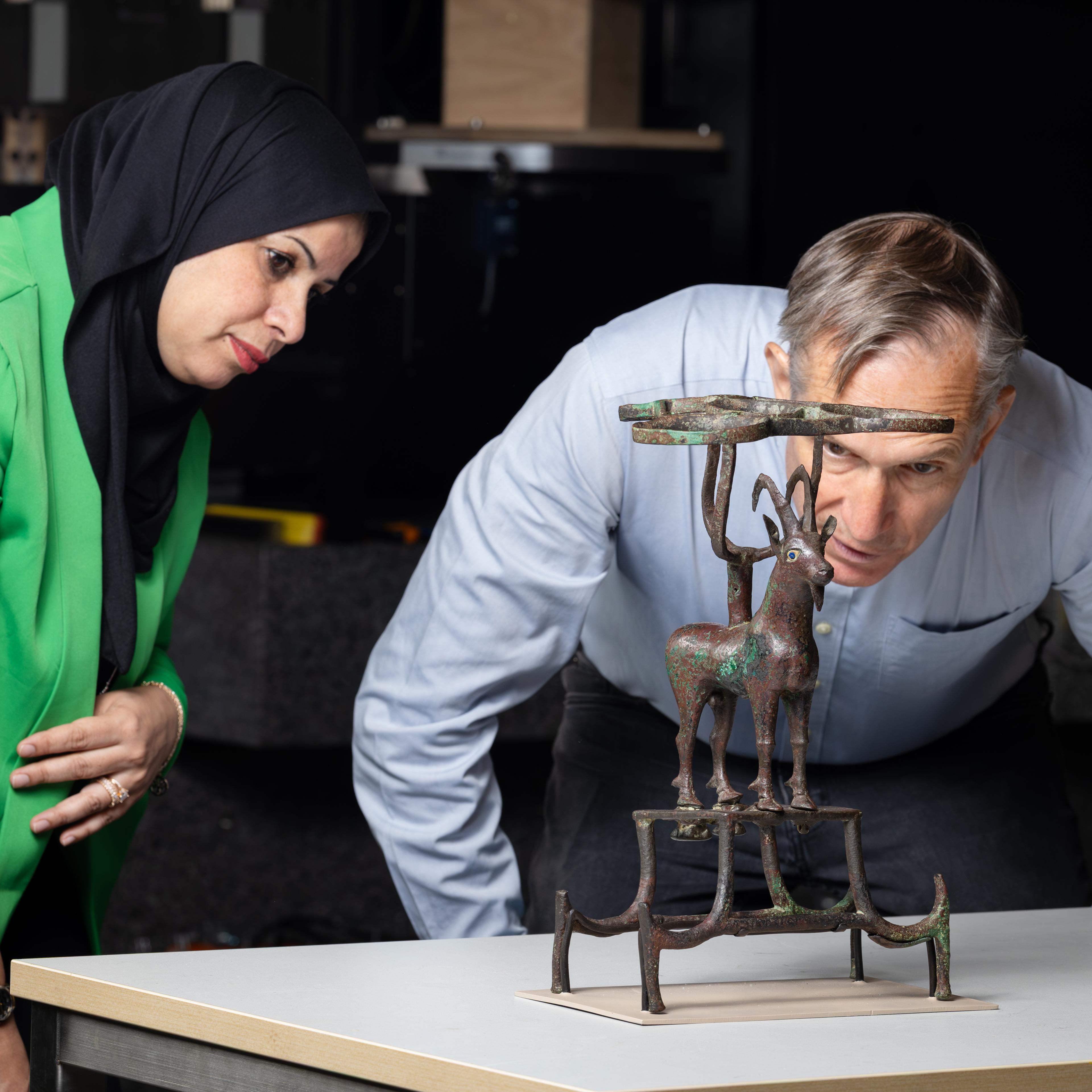 a woman in a green blazer and black headscarf and a man in a blue shirt observing an ibex sculpture