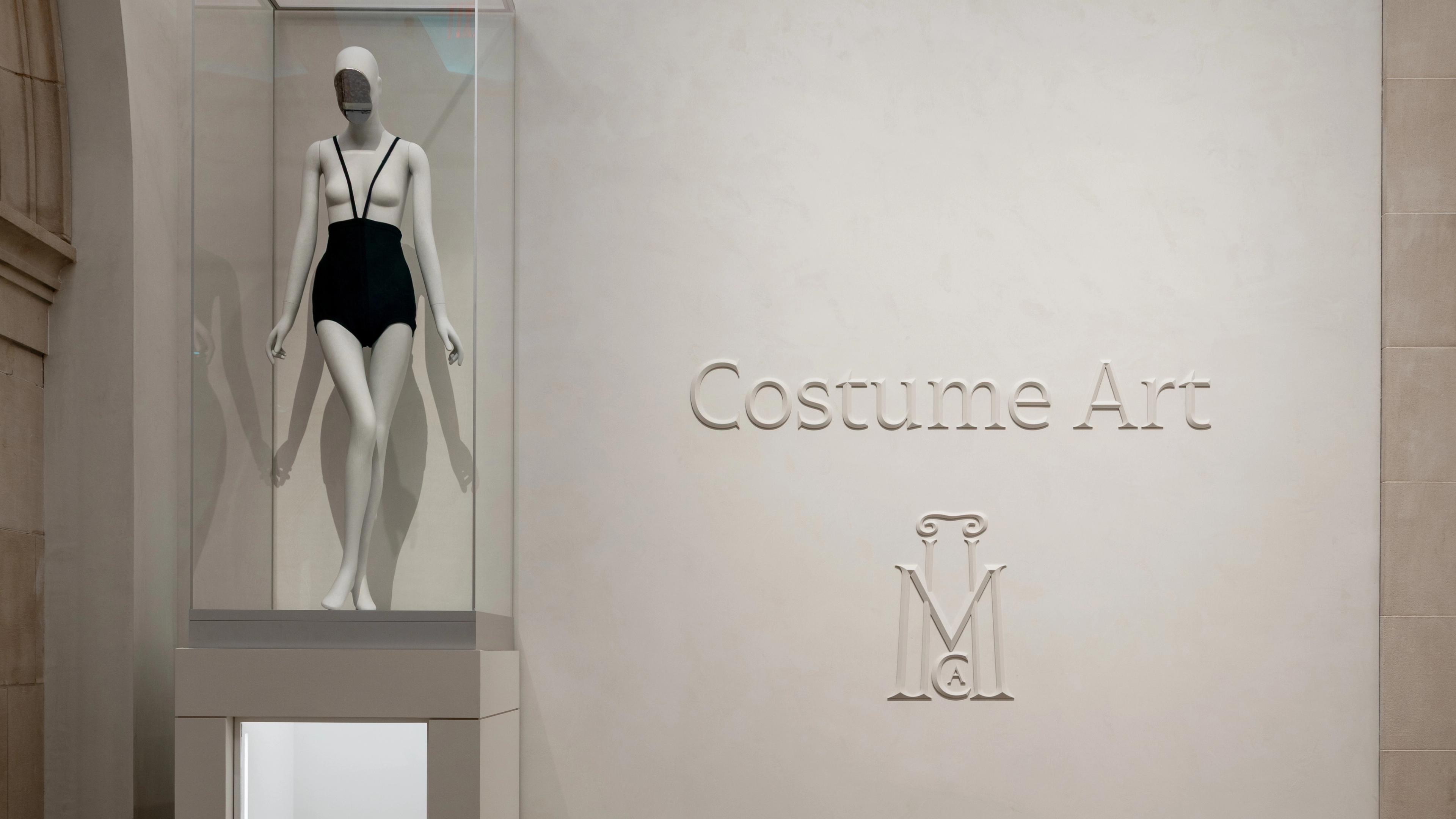 A mannequin wearing a black, strappy swimsuit is displayed in a glass case. Beside it, the wall reads "Costume Art" with an ornate "M" logo below.