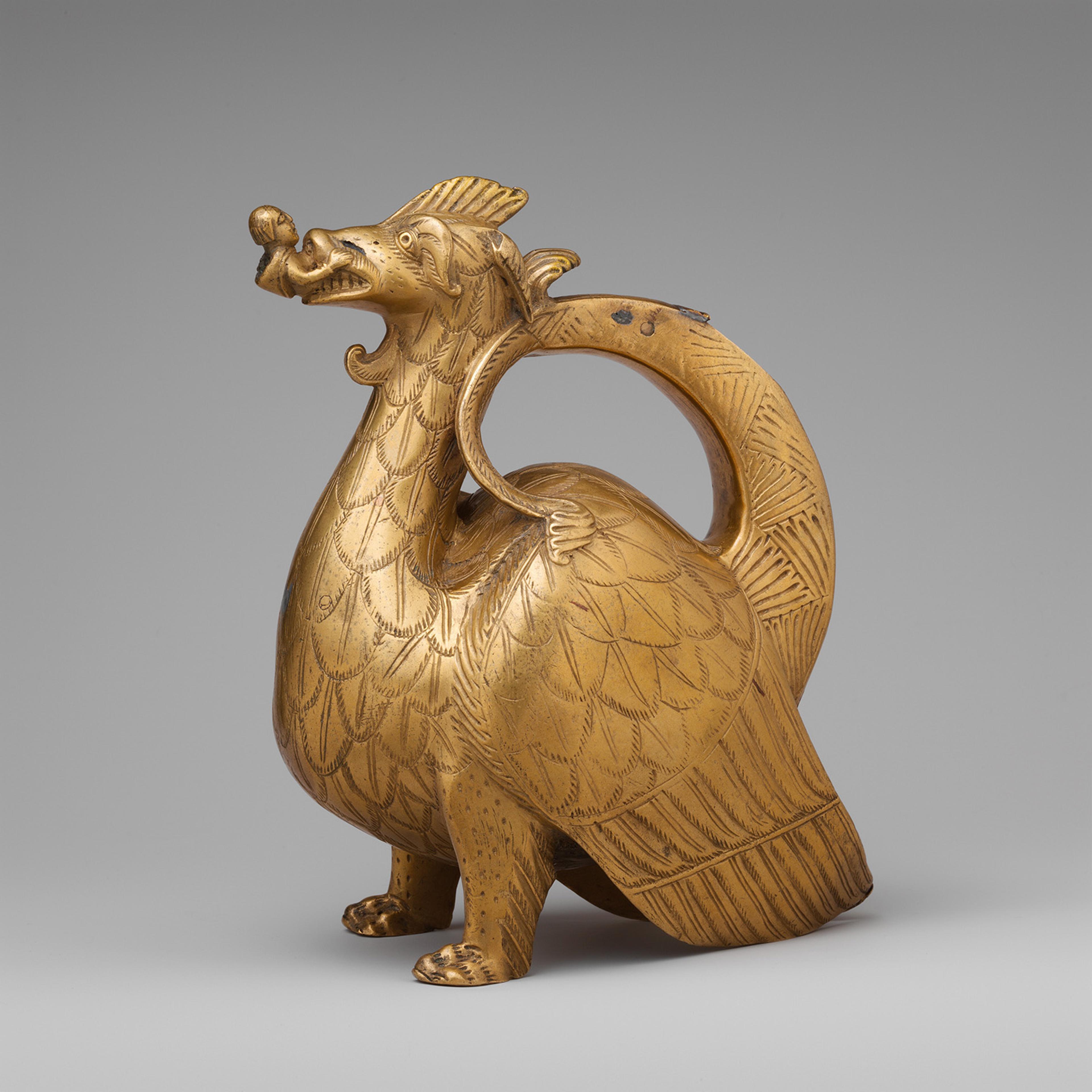 Golden pitcher shaped like a dragon with intricate engraved scales and fierce expression. The handle forms a loop at the back.