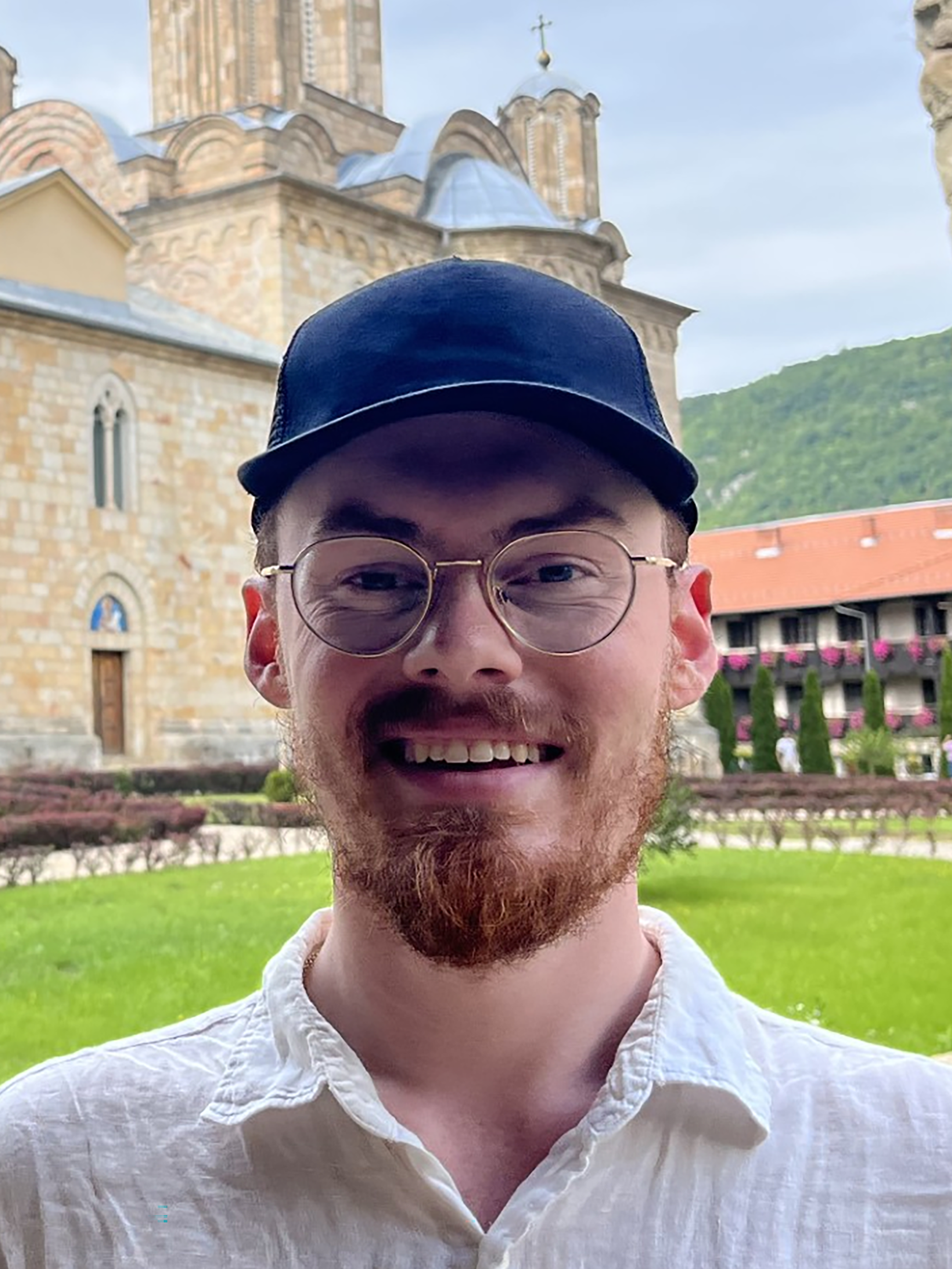 A man wearing glasses and a hat stands in front of a medieval church
