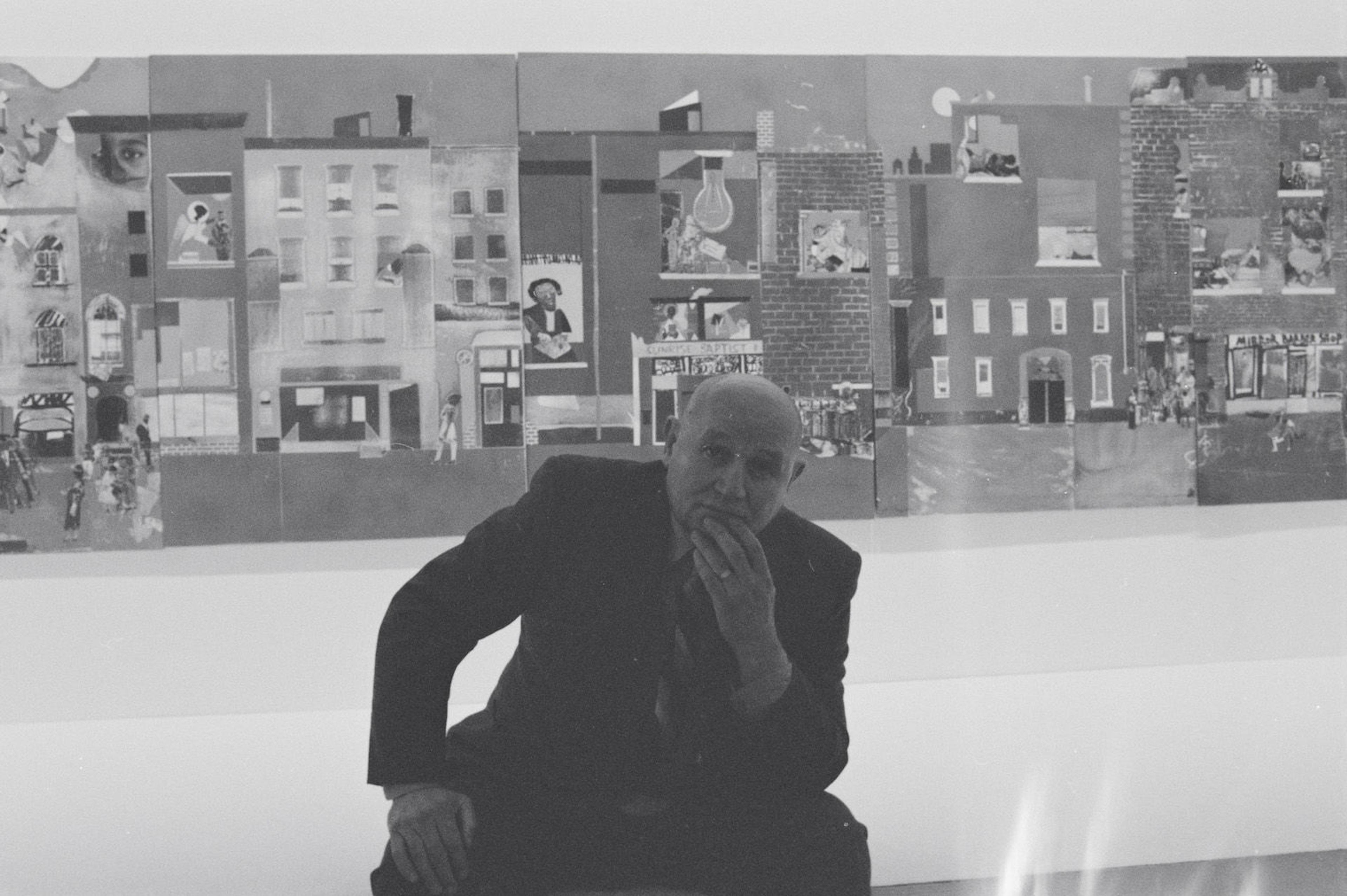 Picture of the artist romare bearden infront of his large paper collage the block. a black and white photograph, bearden is a bald light skinned african american man wearing a suit with a stripped tie.