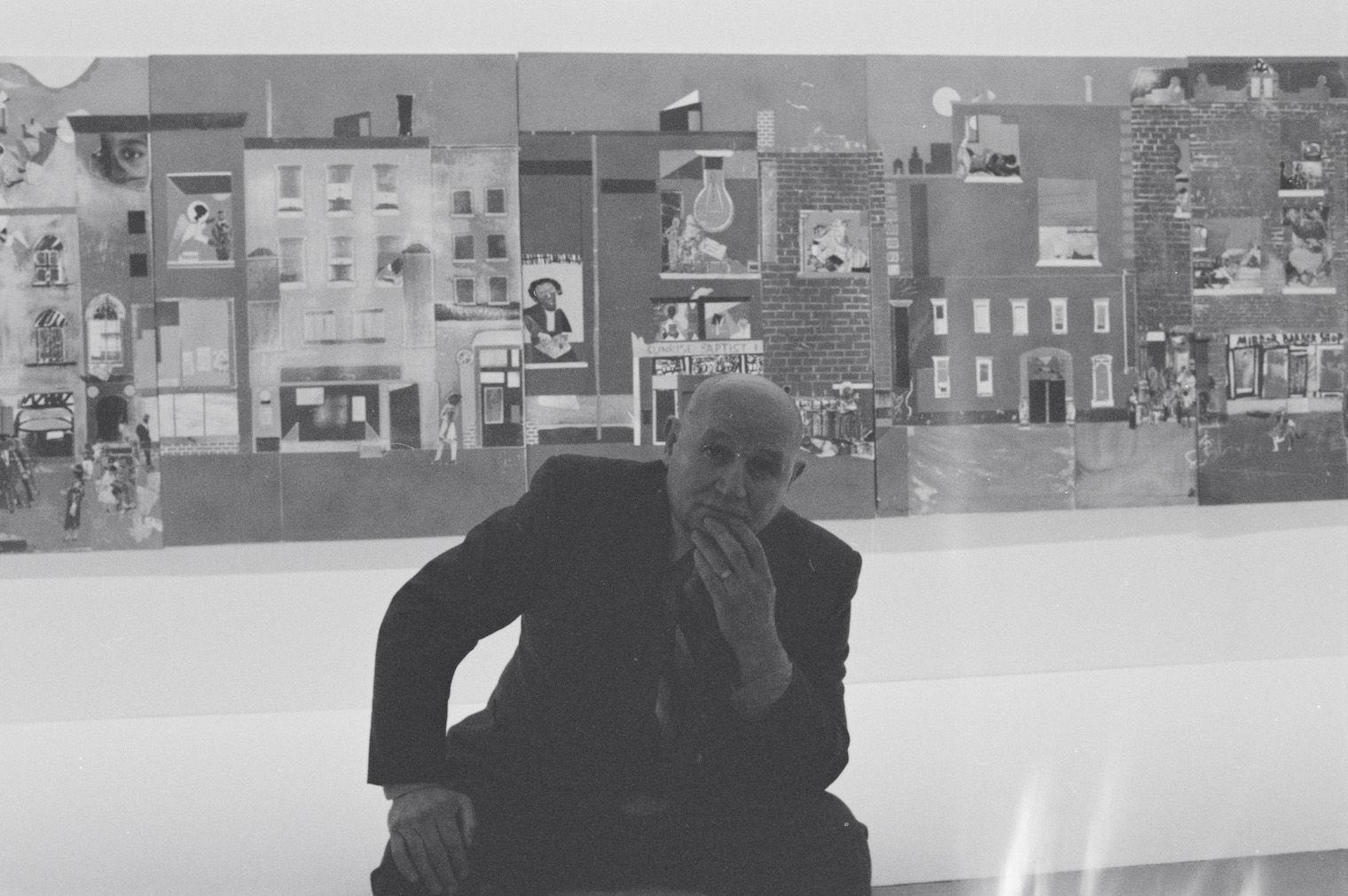 Picture of the artist romare bearden infront of his large paper collage the block. a black and white photograph, bearden is a bald light skinned african american man wearing a suit with a stripped tie.