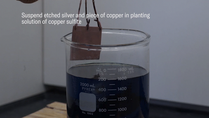 A video animation of an artisan suspending a piece of copper and etched silver in a plating solution of copper sulfite. The artisan attaches a power source to the copper (+) and silver silver (-) and applies an electrical charge. The positively charged ions are plated onto the negatively shared silver not covered by the resist.