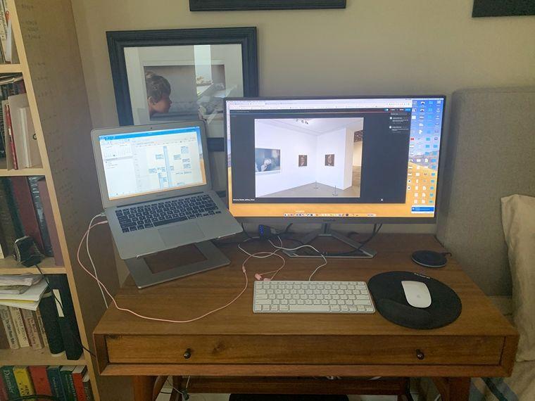 A laptop and monitor are set up with a keyboard and mouse on a desk in a private home, beside a bookshelf and a bed.