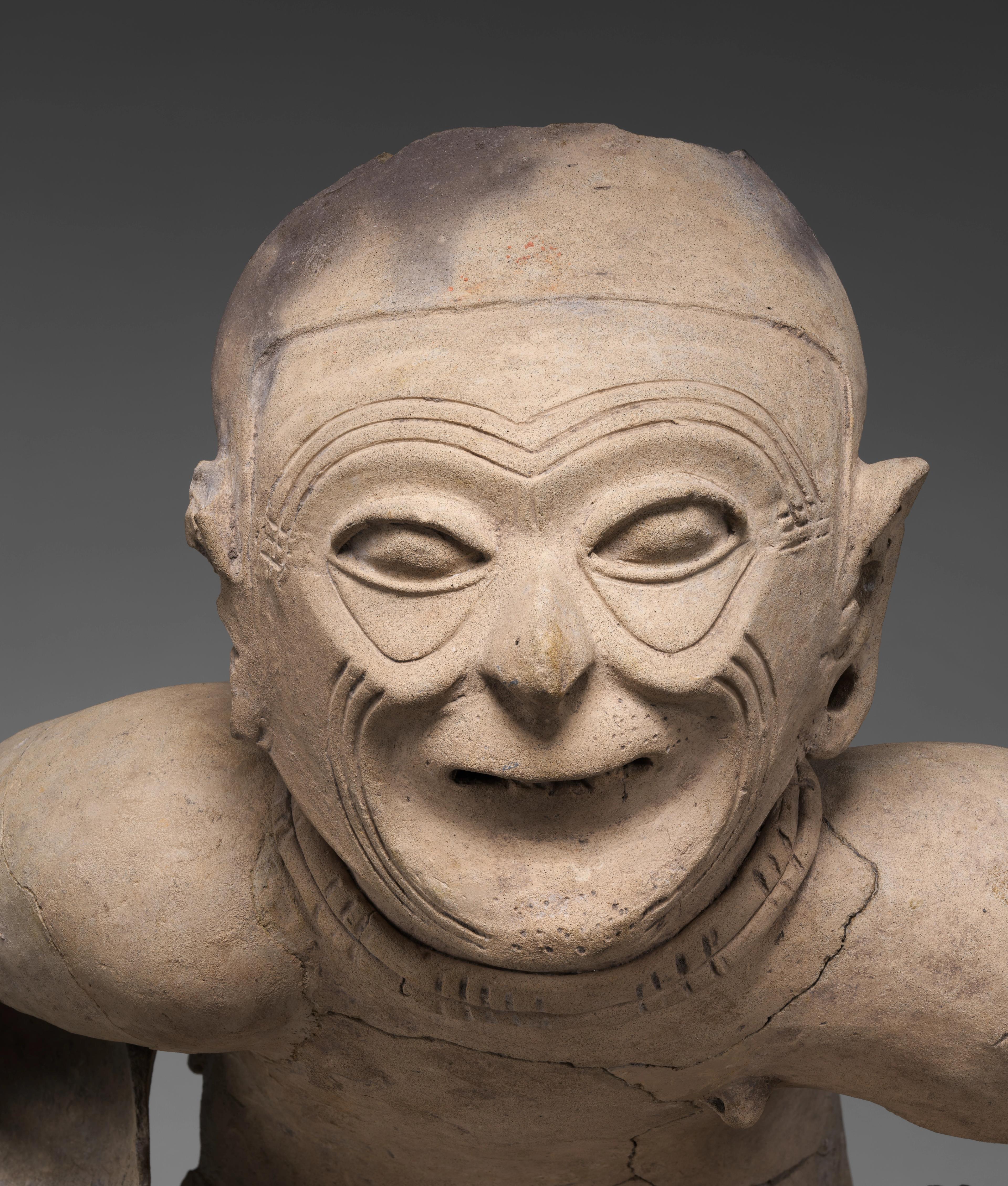 A close-up on the face of a seated elder statue, showing carved wrinkles, protruding eyes, and missing teeth.