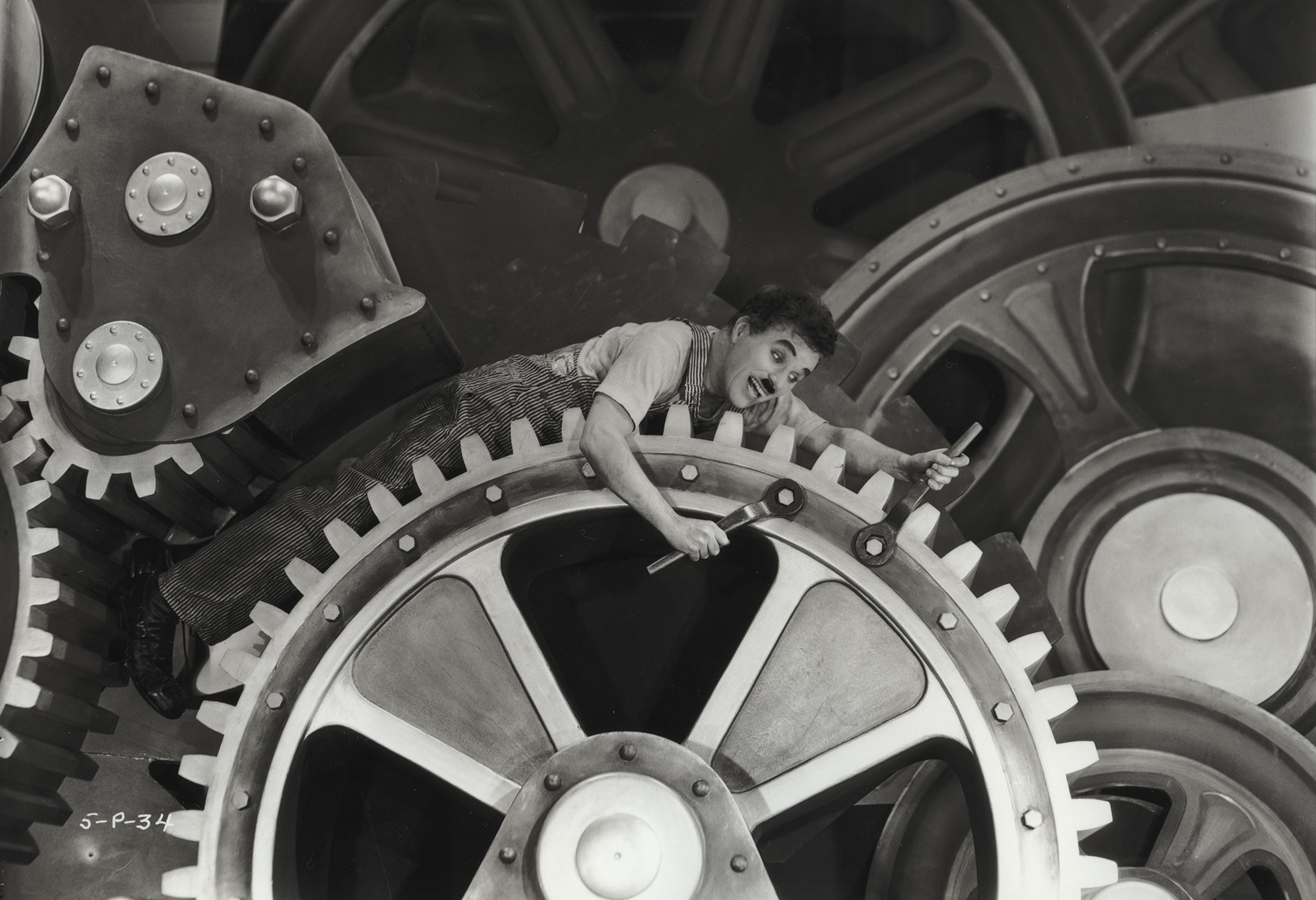 Black and white image of the actor Charlie Chaplin dressed as the tramp, a white man in overalls in this scenario and a mustache where he is trapped between massive factory cogs that he is riding while also trying to tighten the bolts with both of his hands at the same time.