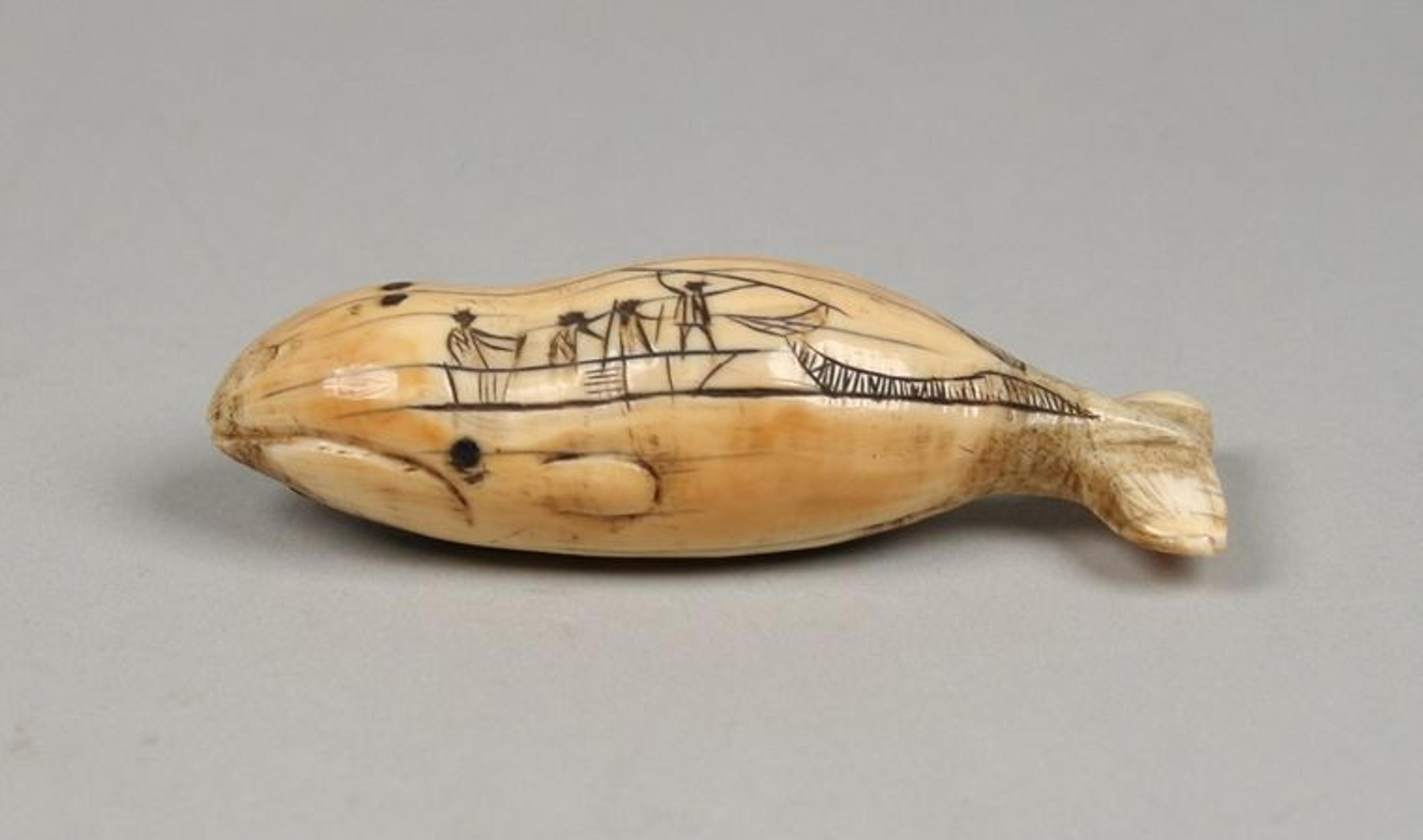 A simple scene of hunters on a boat with the leftmost hunter holding a spear over a whale is etched into a  small piece of ivory that has been carved into the shape of a whale.
