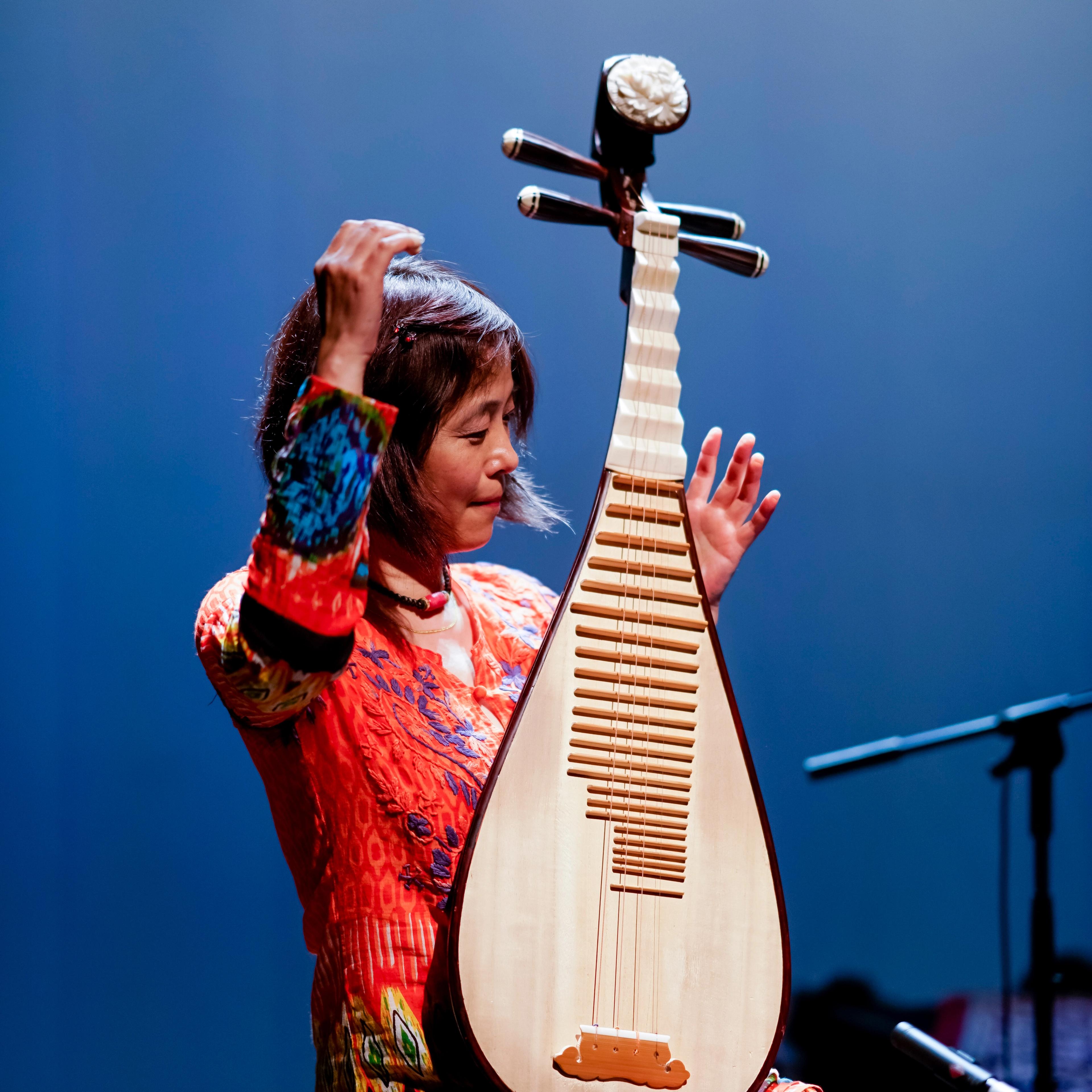 Wu Man, hands aloft, playing the pipa on stage against a deep blue background.