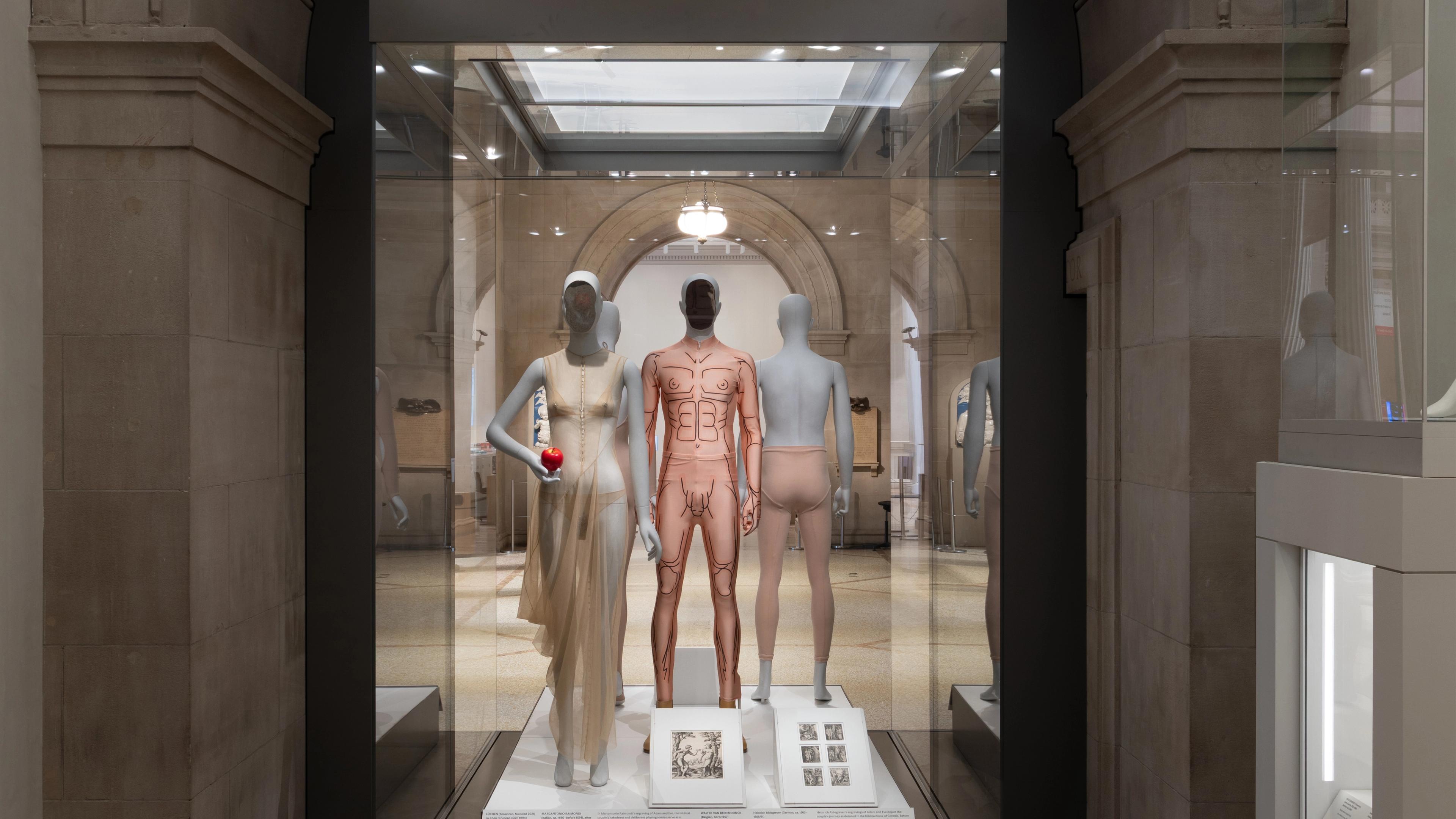 Three mannequins in a museum display showcase fashion designs. One wears a sheer dress, holding an apple. 